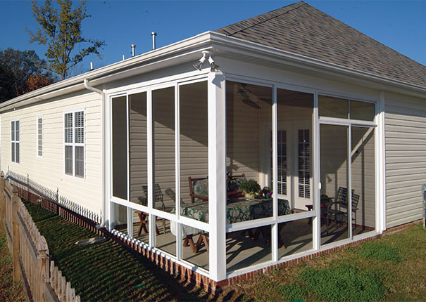A beige house with a corner screened-in porch, white trim, and a fenced backyard on a sunny day.