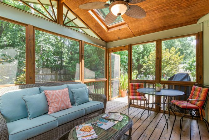 A screened-in porch with a blue sofa, wicker coffee table, dining set, and a ceiling fan under a wood-paneled ceiling.