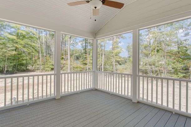 A screened-in porch with gray decking, white railing, and a ceiling fan, overlooking a wooded yard.