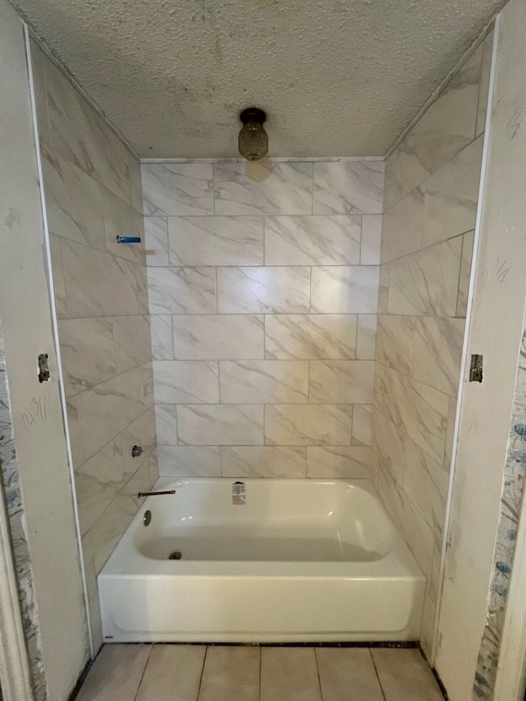 A bathroom with a white bathtub and marble-look tiled walls. An overhead light hangs from the ceiling.