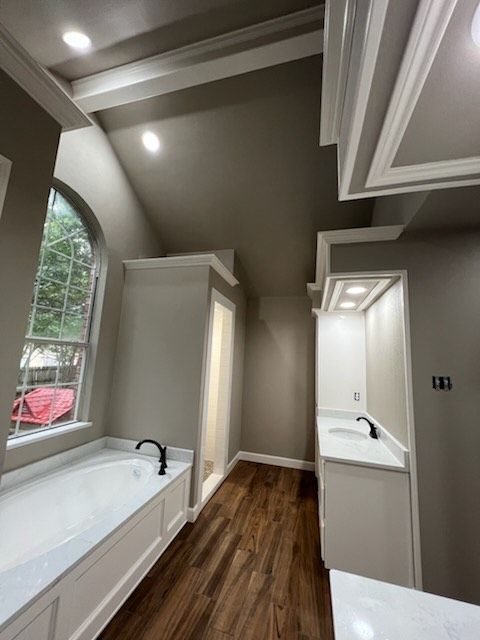 Bathroom with white cabinetry, a bathtub, and a sink. It has a vaulted ceiling, a large arched window, and brown wood flooring.