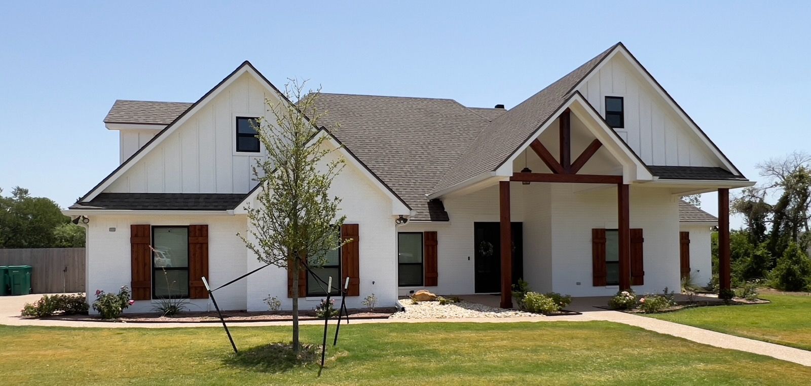 A white, farmhouse-style house with brown shutters and accents. It has a dark roof, with a small tree in the front yard under a clear blue sky.