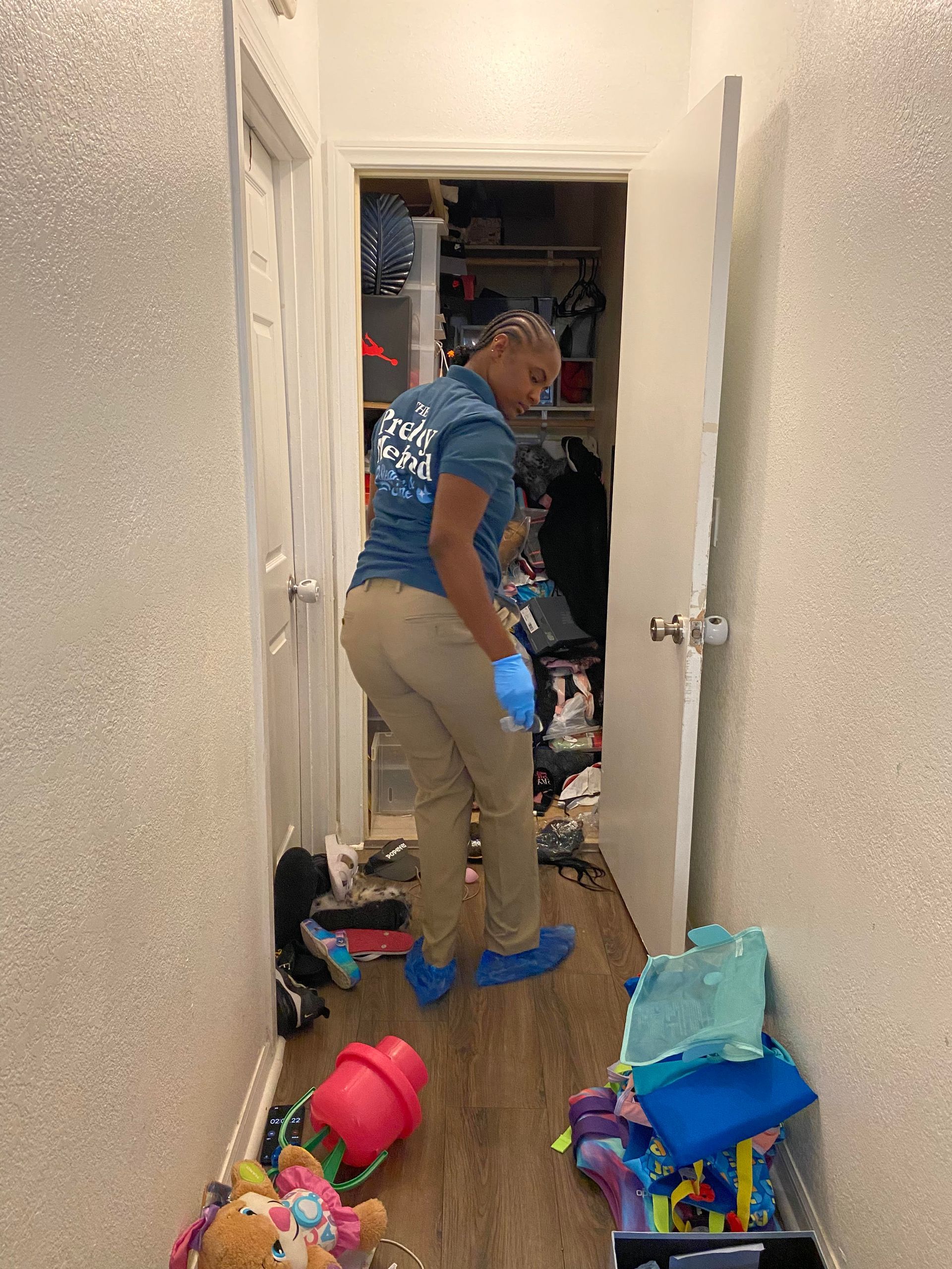A person wearing a blue shirt and gloves stands in a cluttered hallway, looking toward a messy closet.