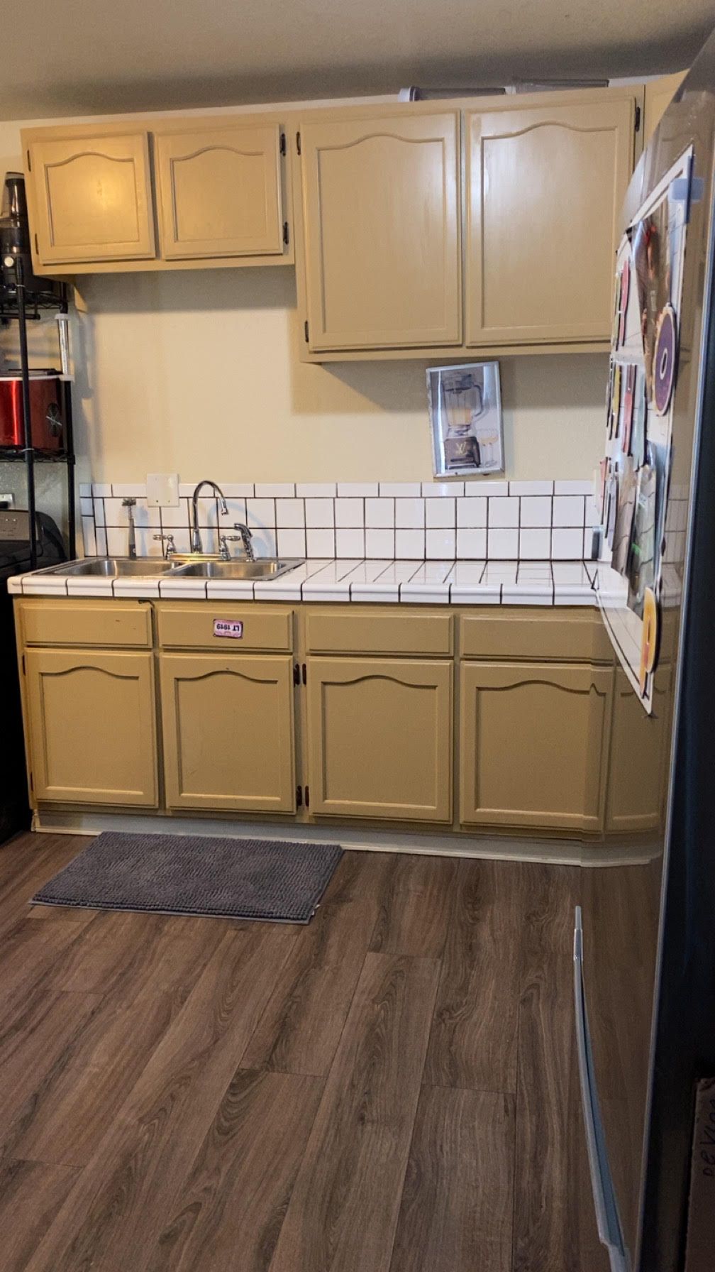 A kitchen with tan cabinets, white tiled backsplash, and a stainless steel sink. The floor is a wood-like laminate.