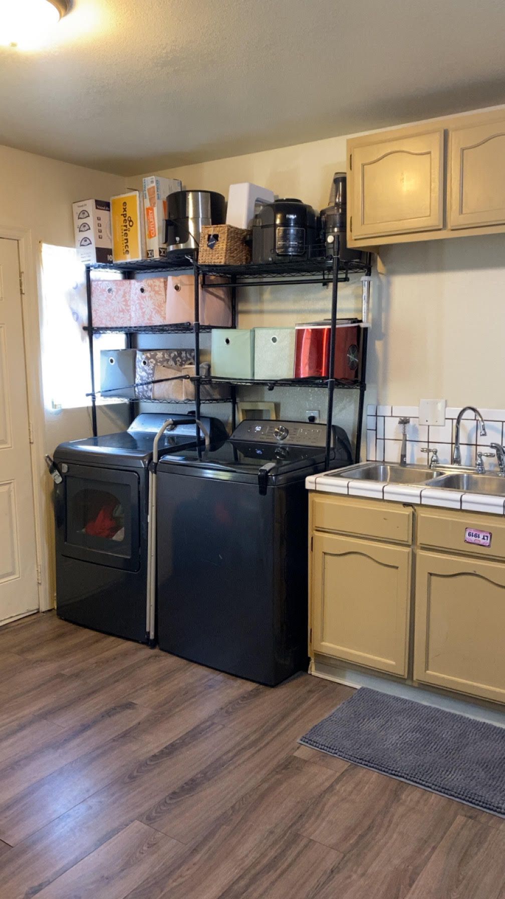 A kitchen with a black washer and dryer, shelves of boxes, and a sink. Beige cabinets and wooden floor.