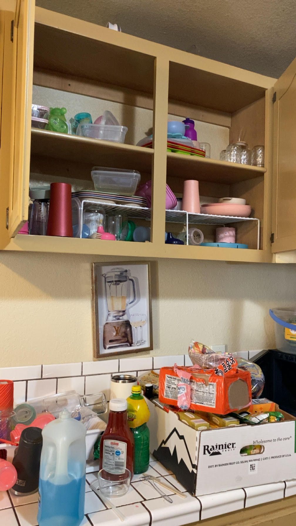 Open kitchen cabinet filled with dishes and cups. Counter has cleaning supplies and boxes of food.