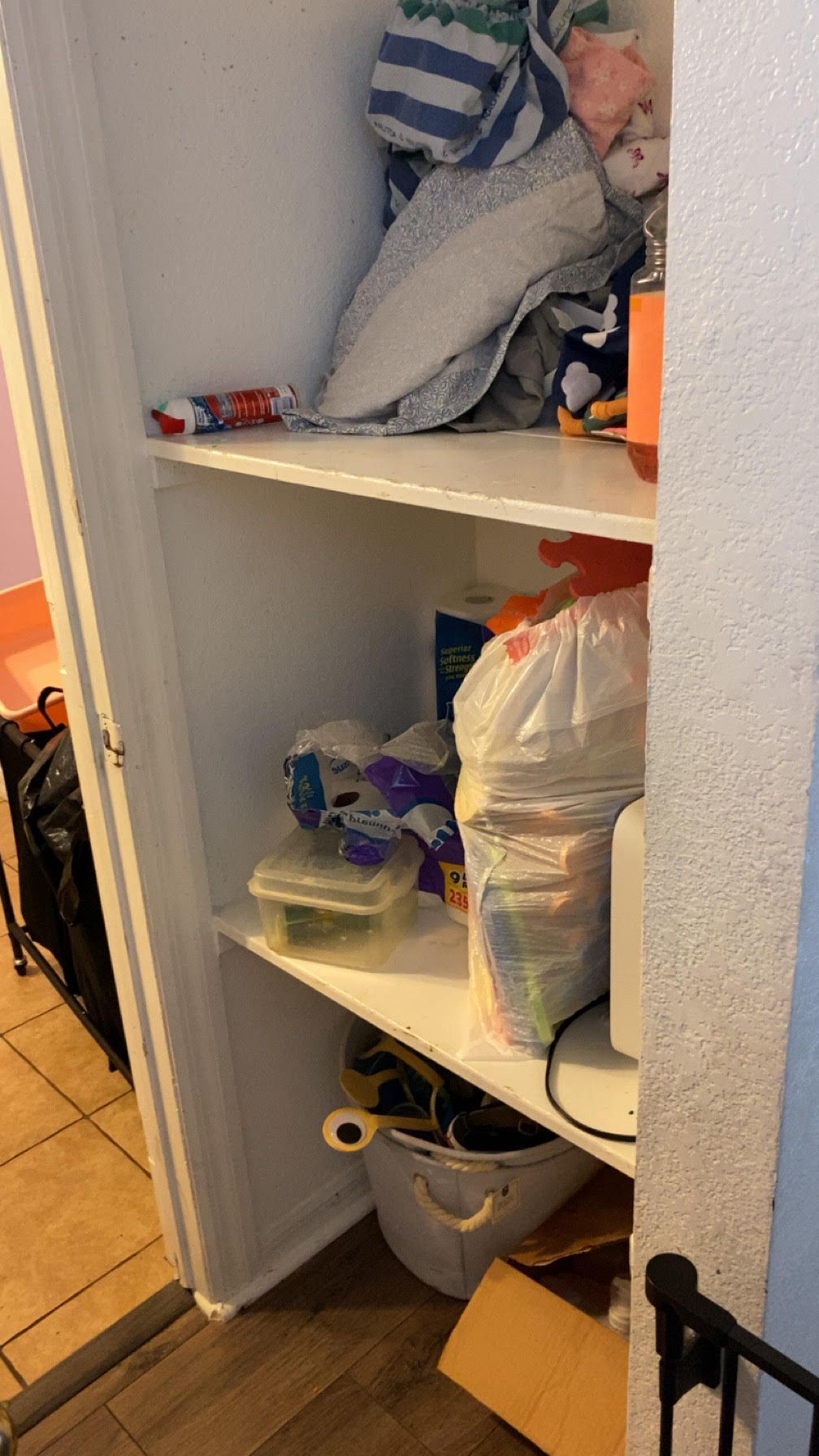White closet with cluttered shelves, containing linens, trash bags, and other household items.