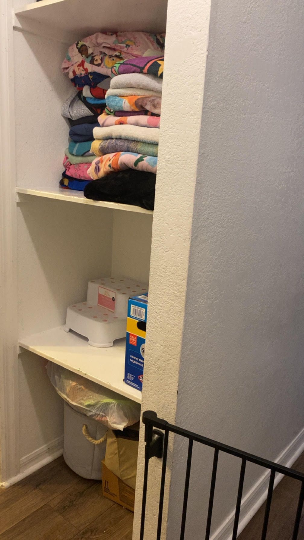 A closet with stacked towels, wipes, and a step stool on shelves. A baby gate and wooden floor are in view.