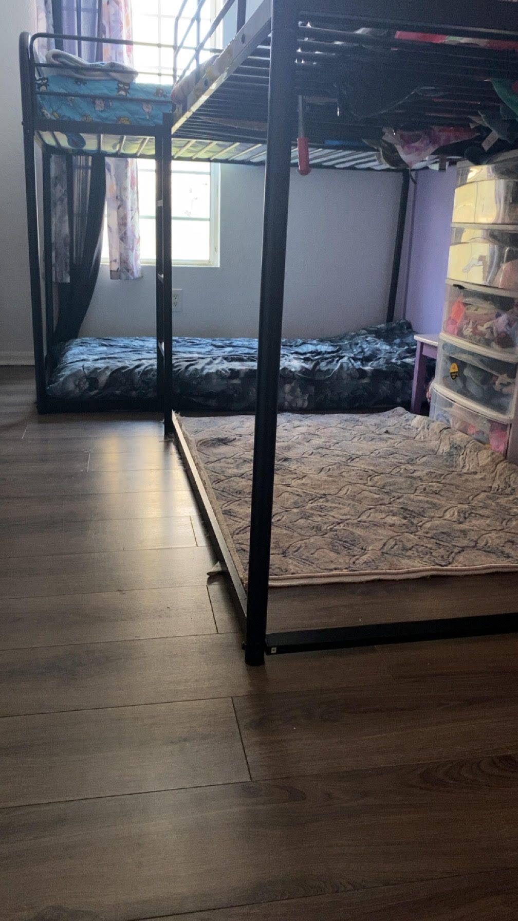 Bunk bed in a bedroom with wooden flooring, a window, and storage containers. Black metal frame with two beds.