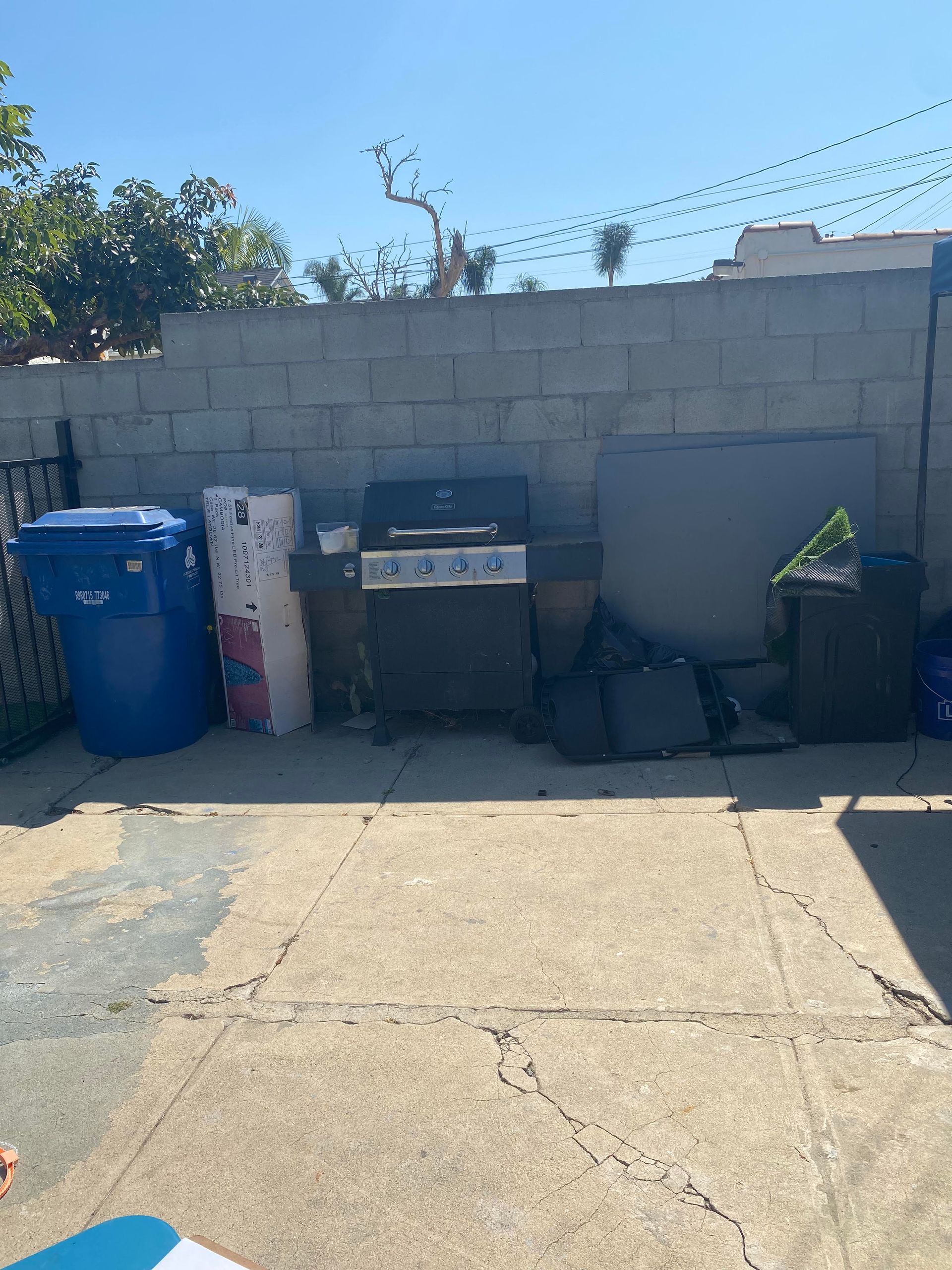 Outdoor scene with a blue trash bin, grill, and various items against a gray brick wall under a sunny sky.