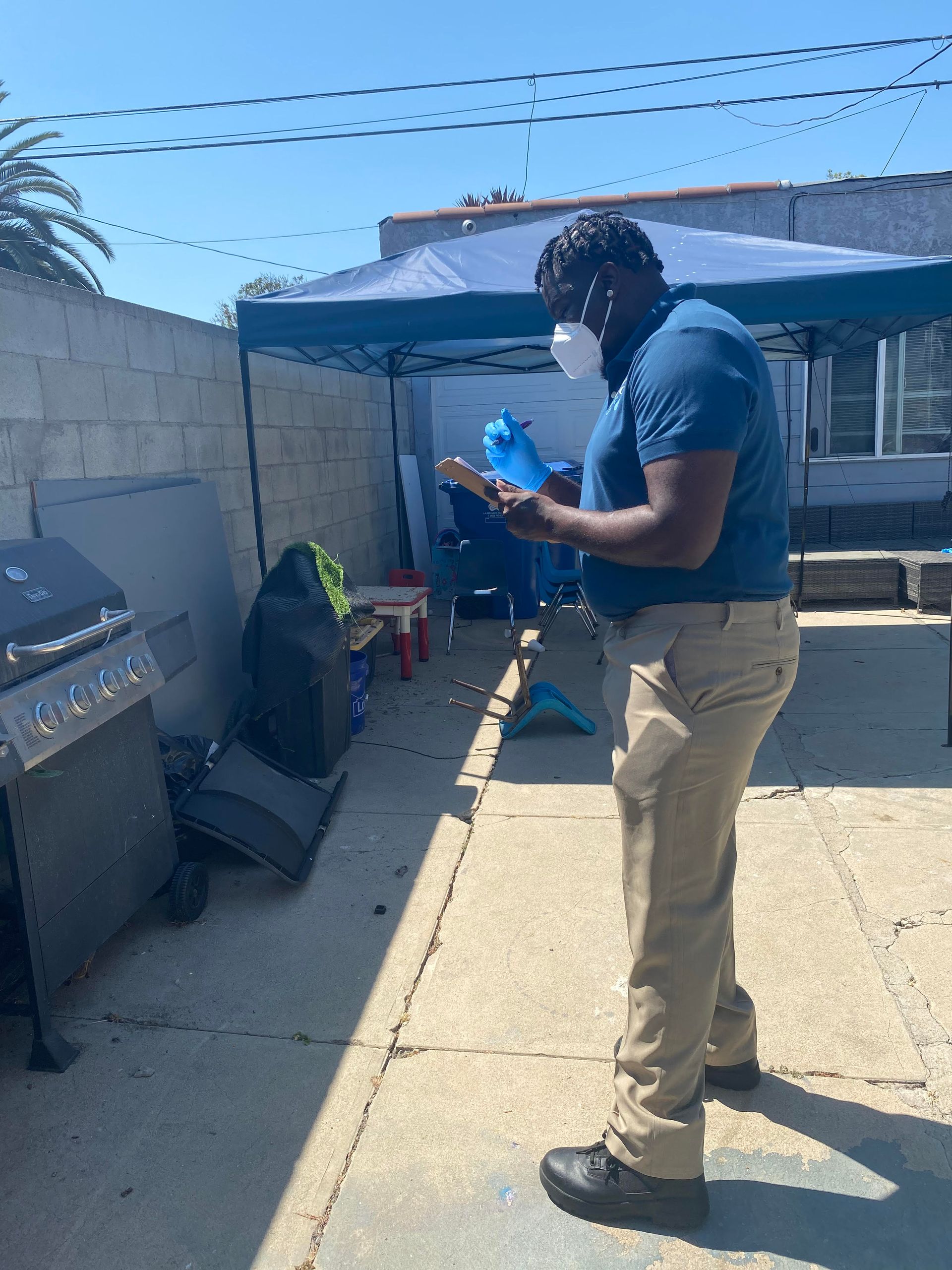 A man wearing a face mask and khaki pants, stands outside and looks at an object. There's a grill and tarp in the background.