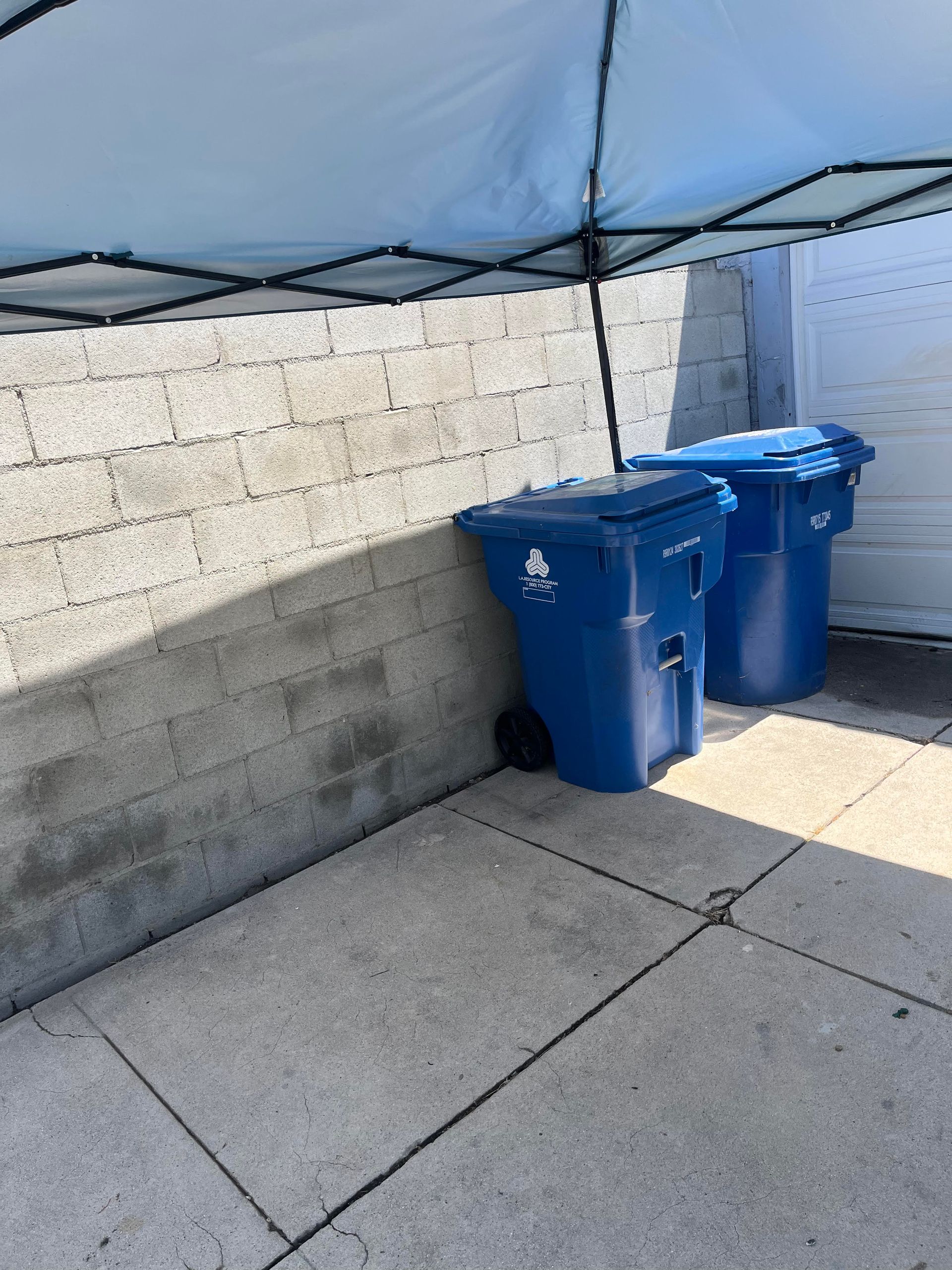Two blue trash bins sit on a concrete surface next to a cinder block wall and a white garage door, under a blue canopy.