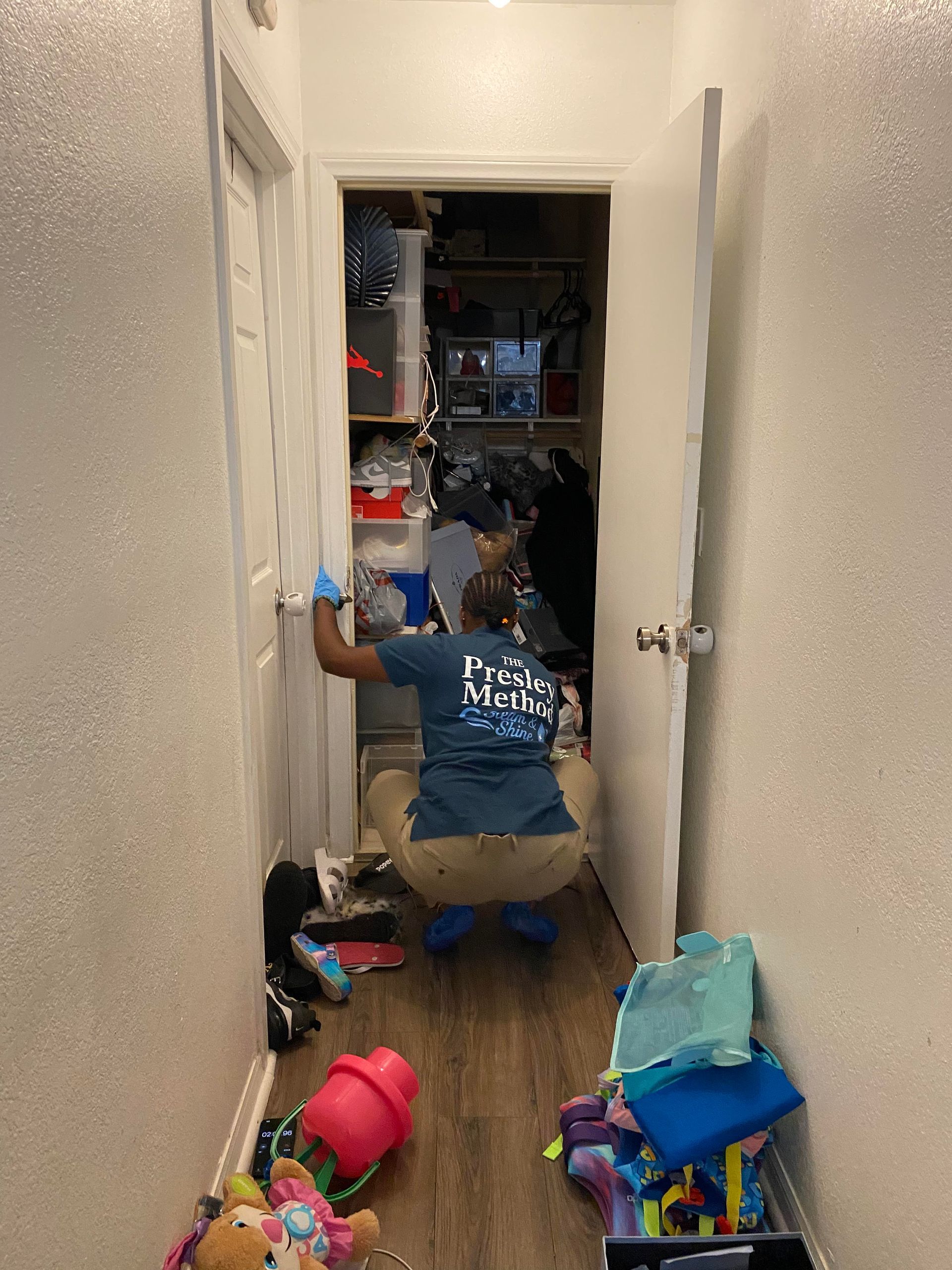 Person cleaning a cluttered closet in a narrow hallway. They are wearing a blue shirt and blue gloves and kneeling.