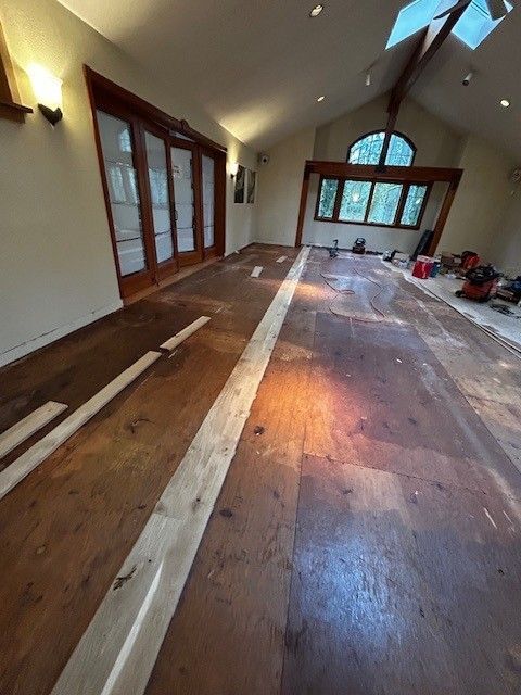 A room under renovation with partially installed hardwood flooring, large windows, and exposed subfloor planks. A room under renovation with partially installed hardwood flooring, large windows, and exposed subfloor planks.