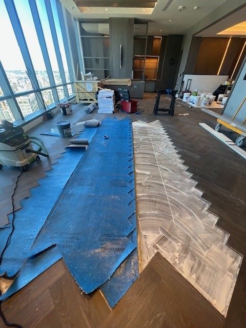 An interior construction site features a room with a herringbone wood floor, blue protective sheeting, and adhesive. An interior construction site features a room with a herringbone wood floor, blue protective sheeting, and adhesive.