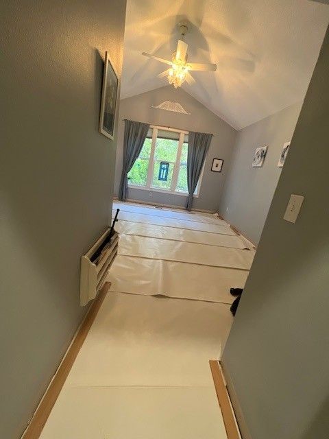 A view down a hallway into a room with gray walls, a ceiling fan, and paper floor protection covering the floor. A view down a hallway into a room with gray walls, a ceiling fan, and paper floor protection covering the floor.