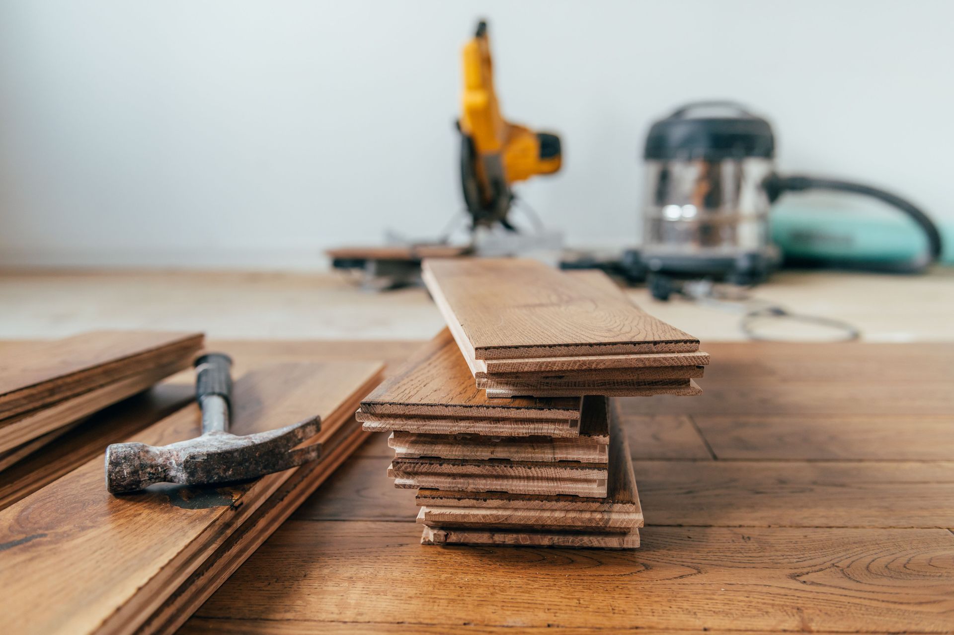 A hammer rests on wood flooring planks next to a stacked pile of flooring, with a power tool and vacuum in the background. A hammer rests on wood flooring planks next to a stacked pile of flooring, with a power tool and vacuum in the background.