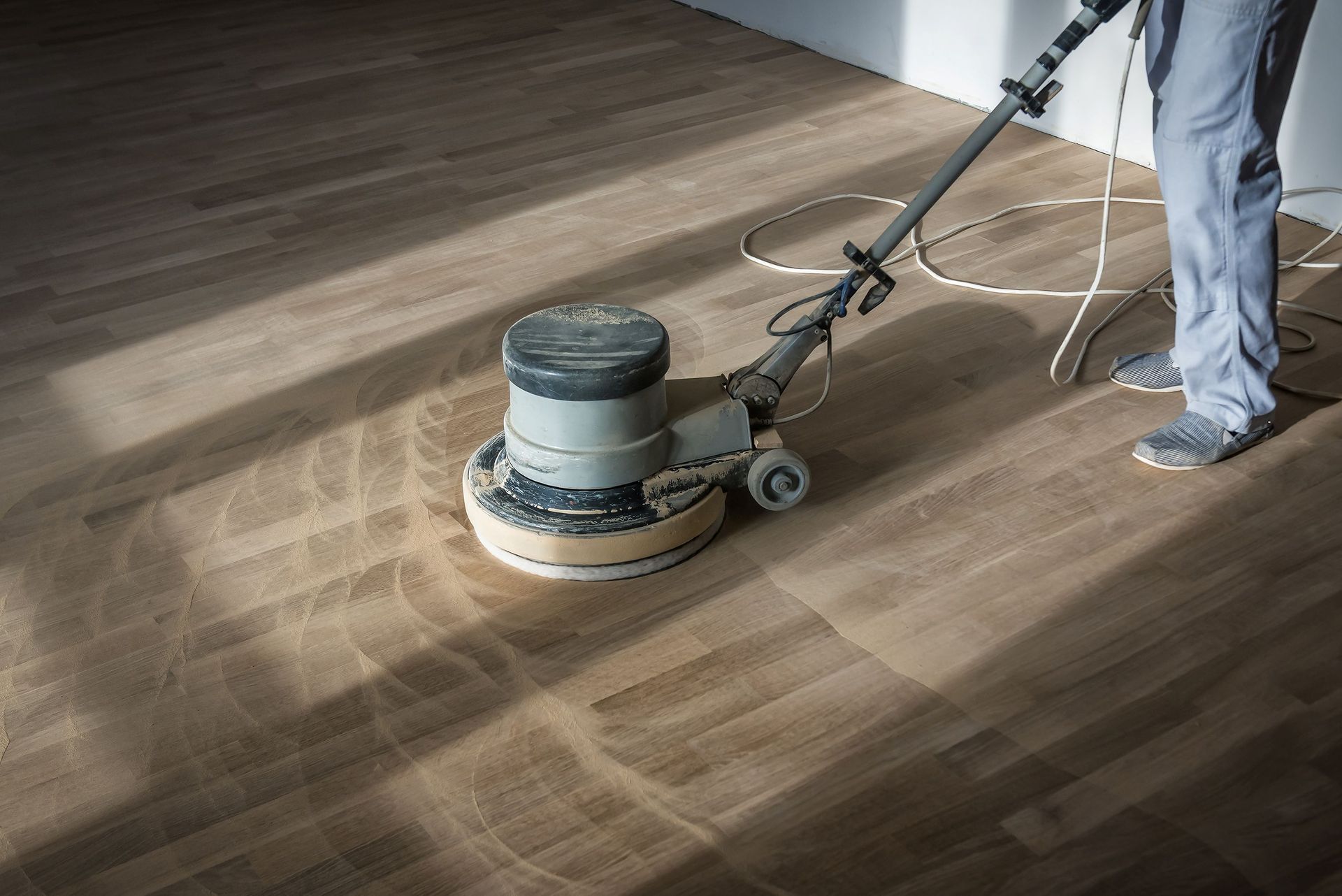 A person uses a floor buffer machine to sand and refinish a light-colored wooden floor. A person uses a floor buffer machine to sand and refinish a light-colored wooden floor.