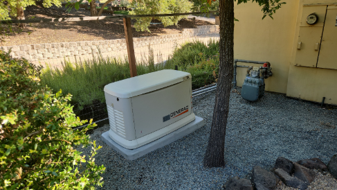 A white generator is sitting under a tree in front of a house.