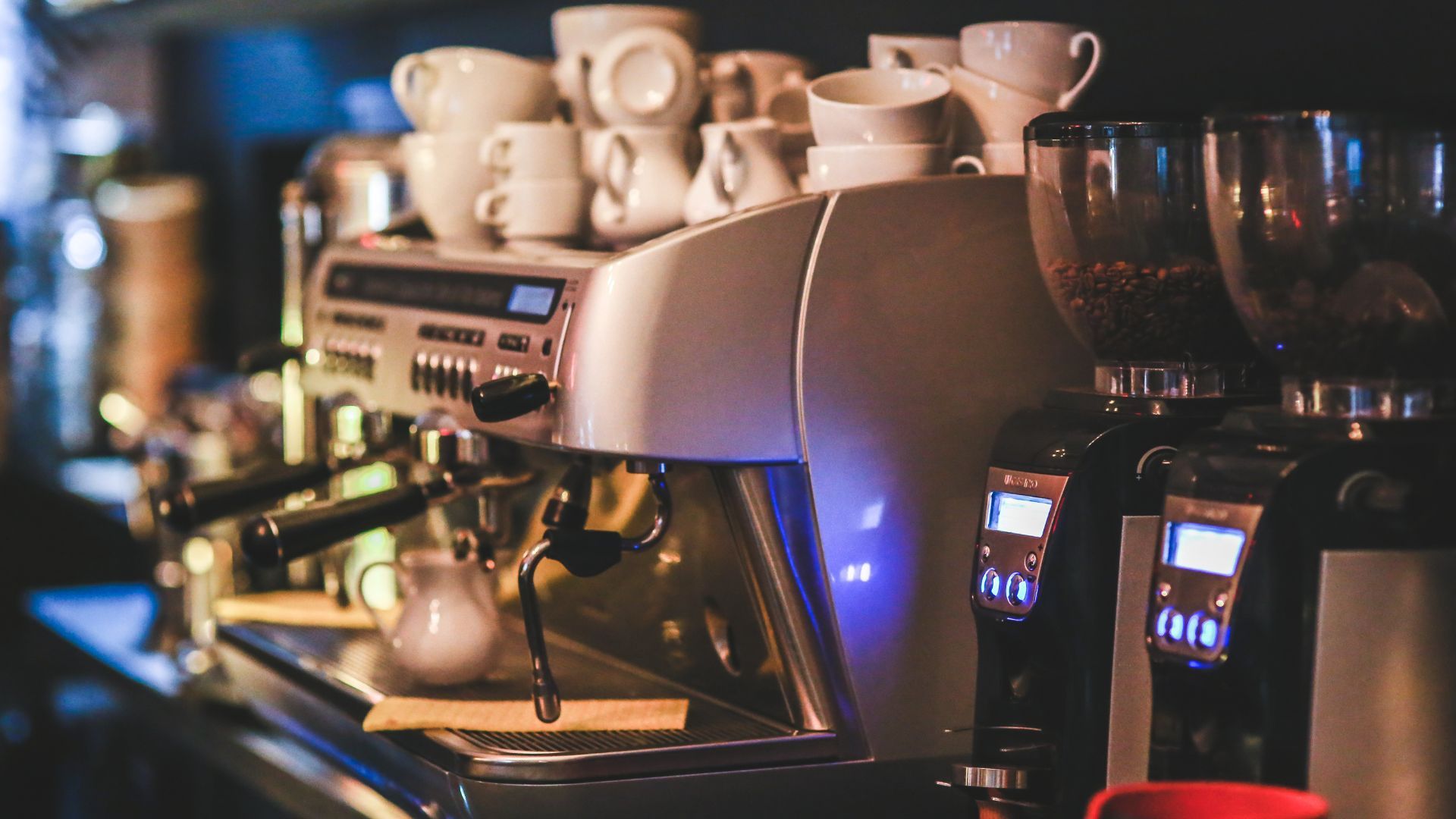 Espresso machine and grinders in a cafe setting, white cups stacked in the background.