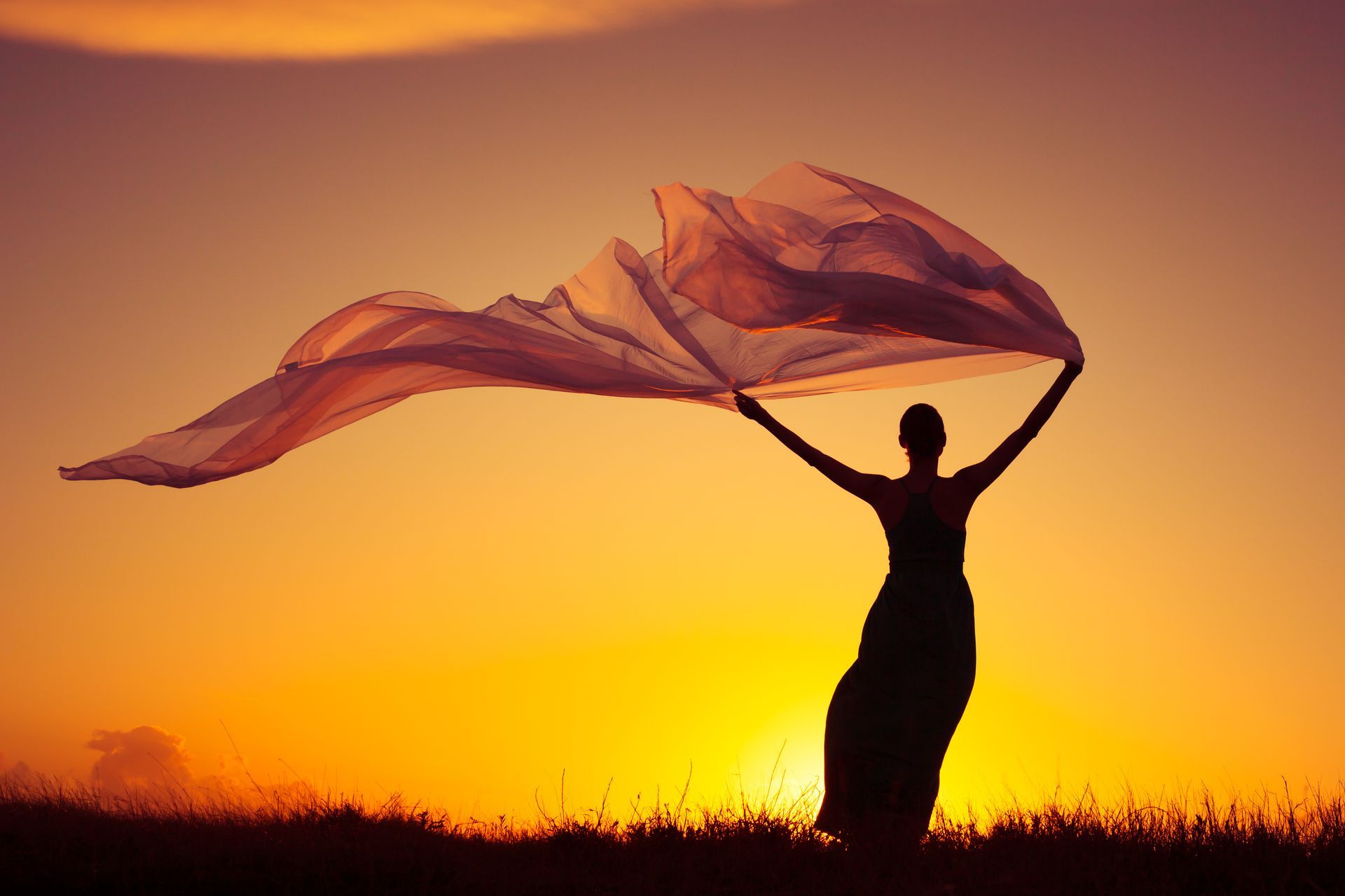 A woman is holding a scarf in the wind at sunset.