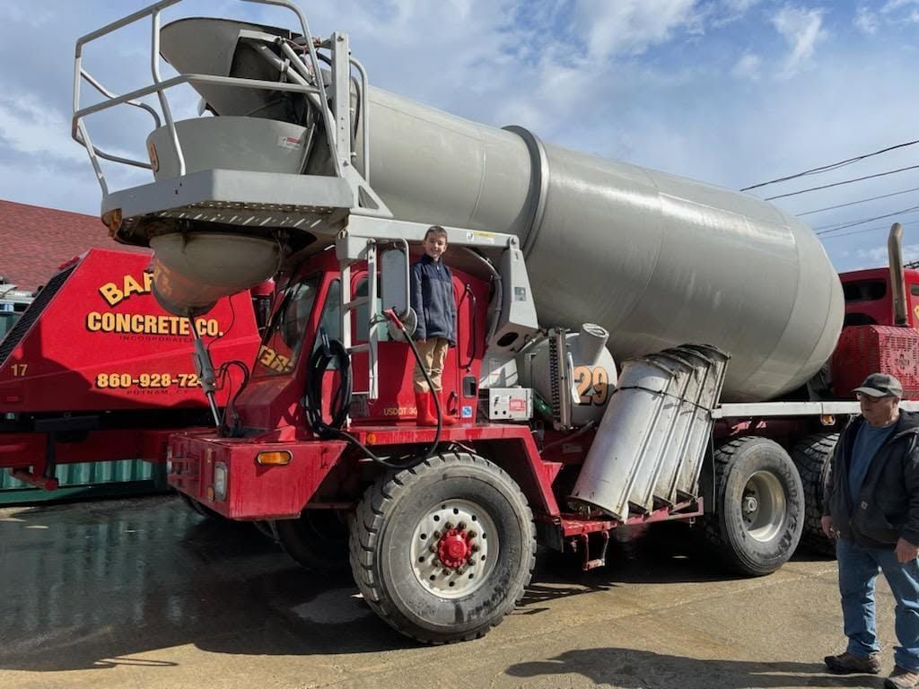 Large red concrete mixer truck with a silver drum parked outdoors, with a man standing beside it.