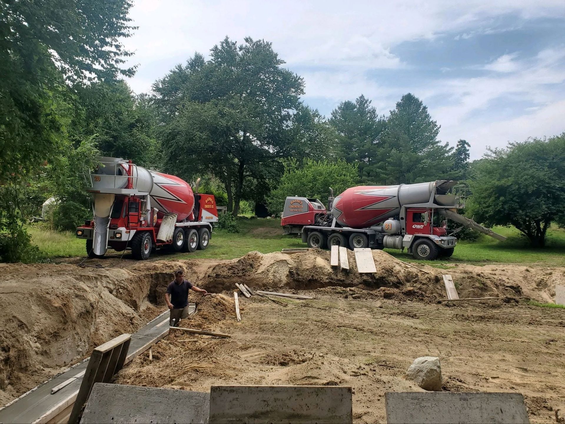 Two red cement mixer trucks beside a large muddy excavation site in a wooded area