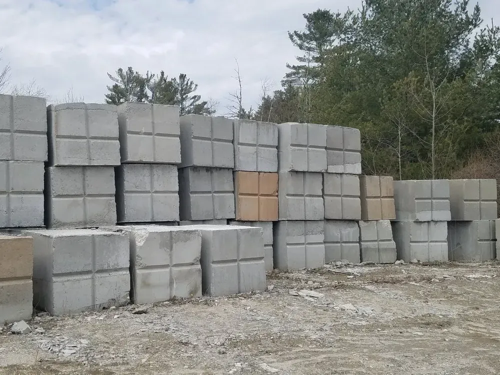 Stacks of gray concrete blocks outdoors beside a gravel lot and trees