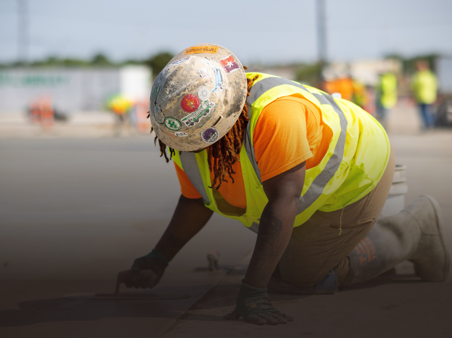 A construction worker wearing a hard hat and safety vest is kneeling on the ground.
