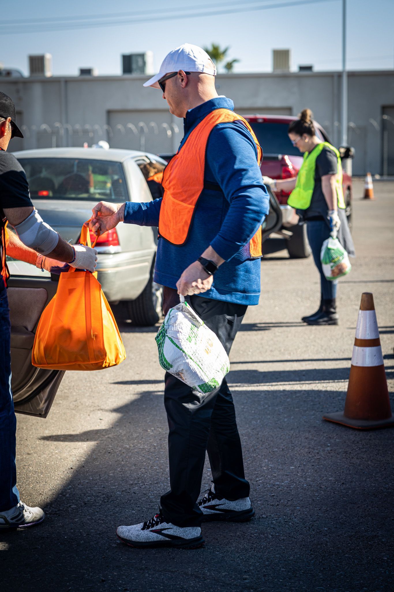 A man in an orange vest is giving a bag of food to another man in a parking lot.