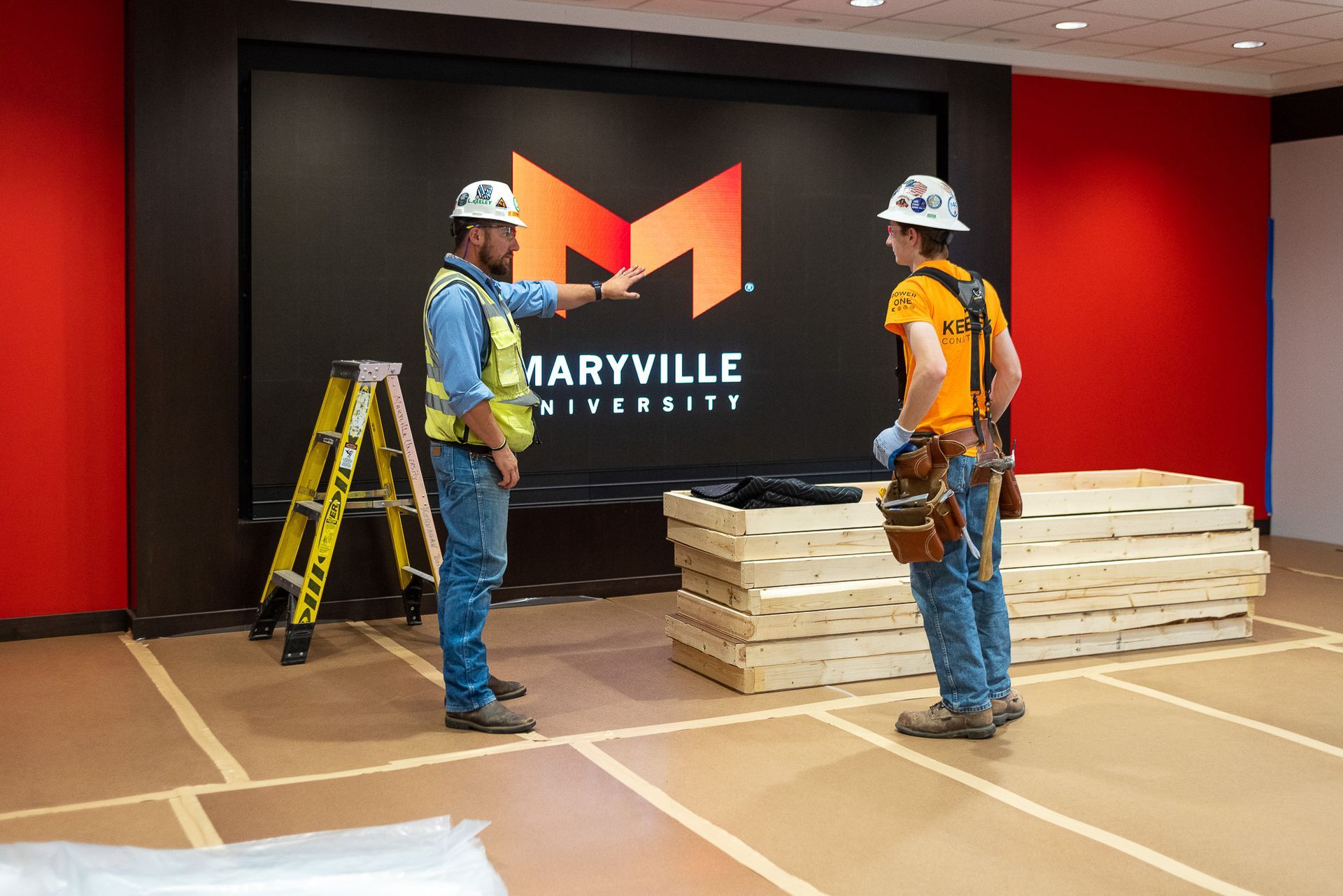 Two construction workers are standing in front of a large sign that says maryville university.