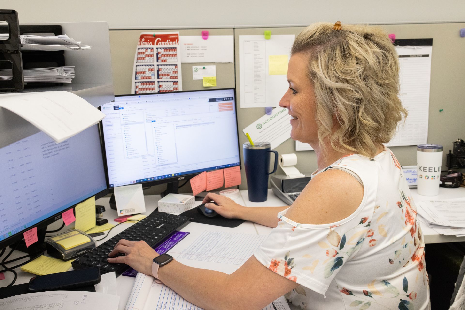 A woman is sitting at a desk in front of two computer monitors.