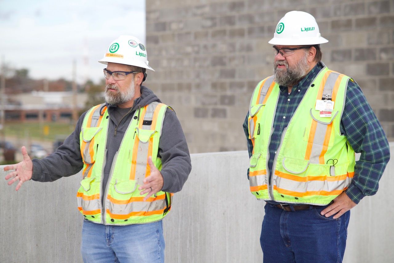 Two construction workers wearing hard hats and safety vests are standing next to each other.
