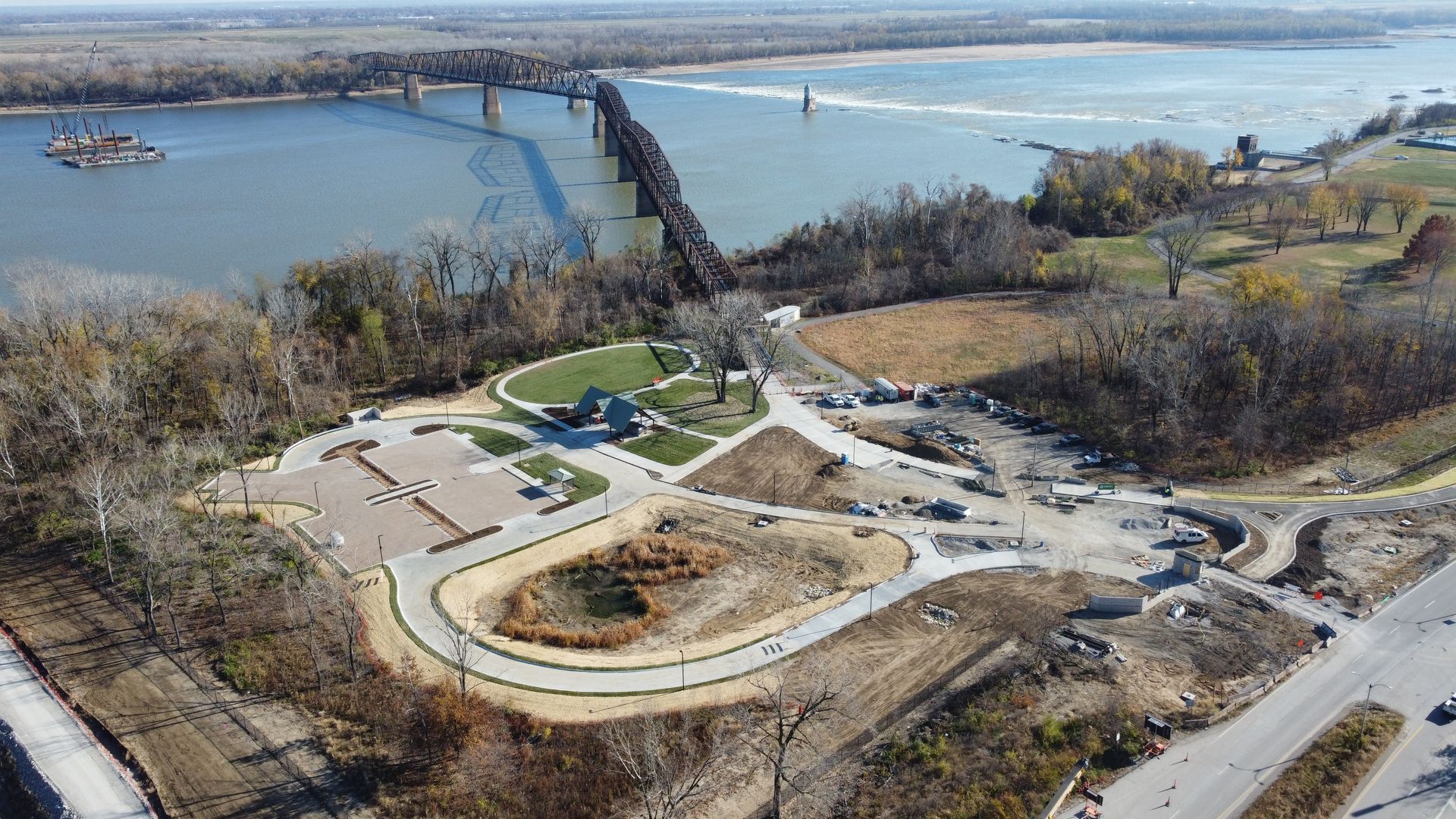 An aerial view of a park next to a river with a bridge in the background.