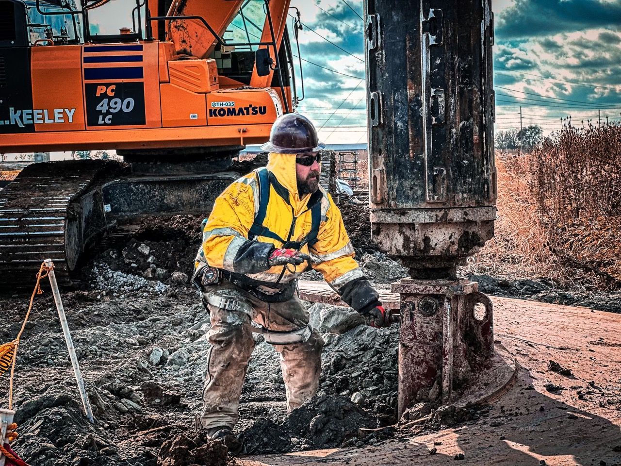 A man in a yellow jacket is working in the dirt in front of a komatsu excavator.