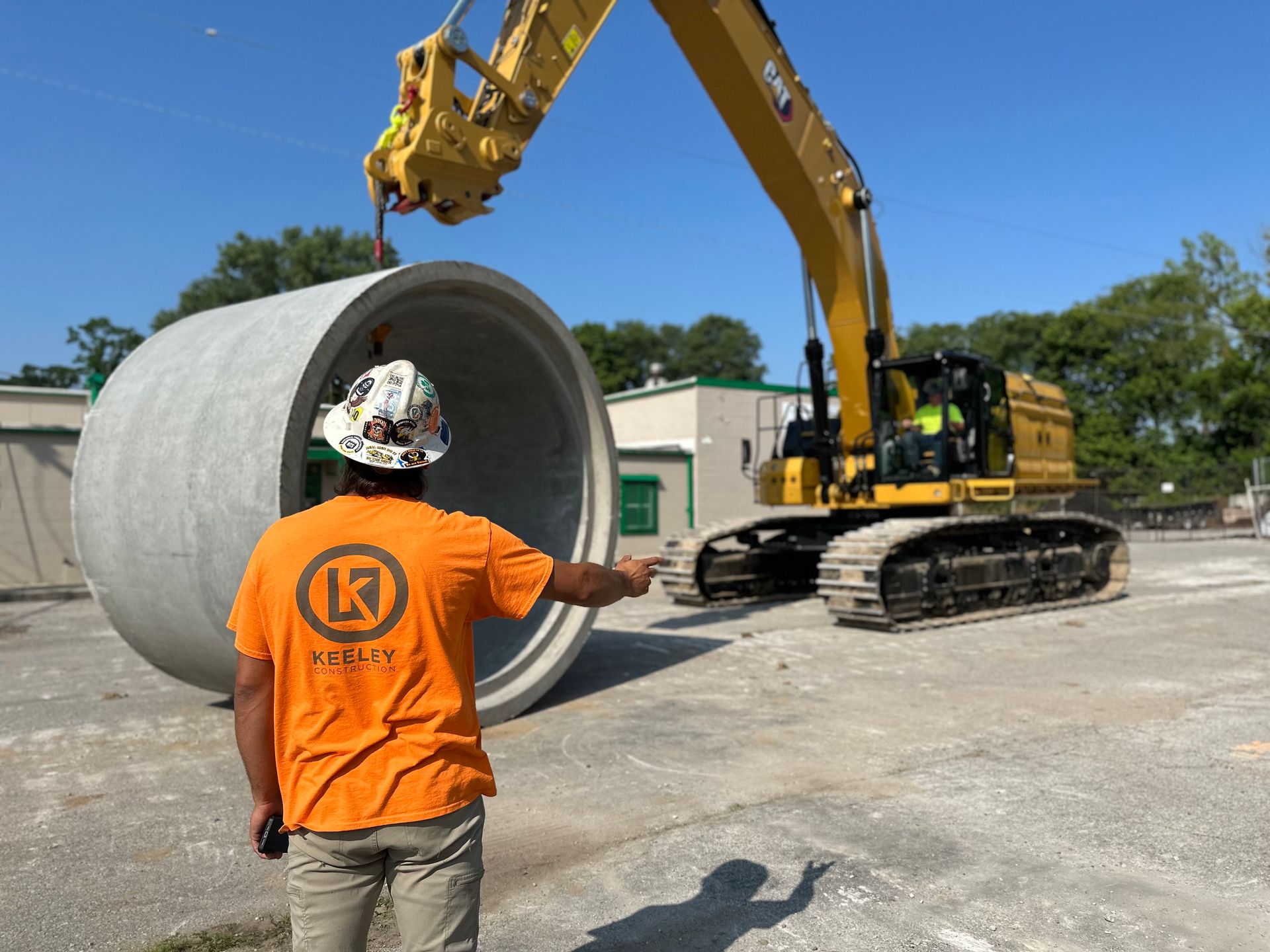 A man in an orange shirt is pointing at a large concrete pipe being lifted by a crane.