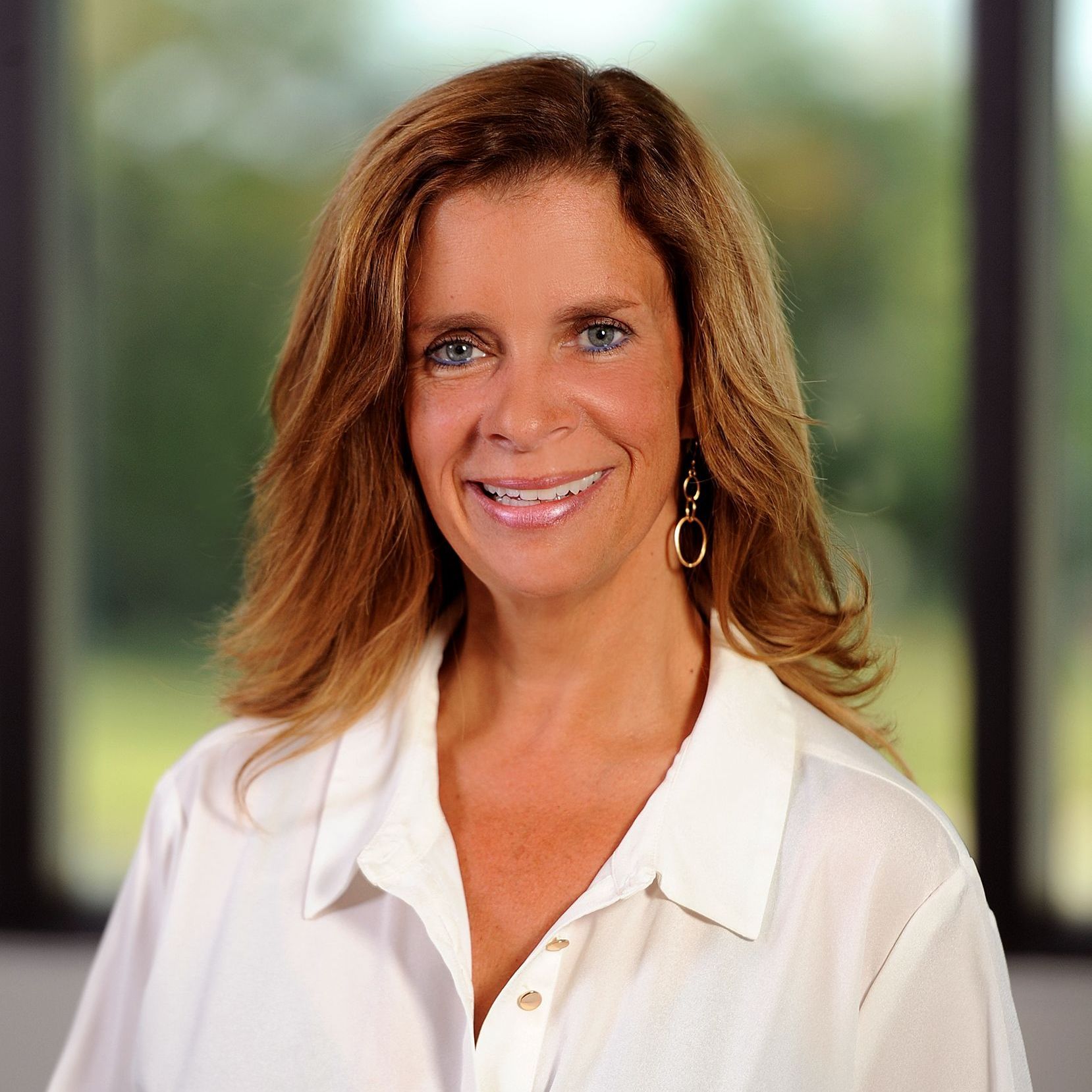 A woman wearing a white shirt and earrings is smiling in front of a window.