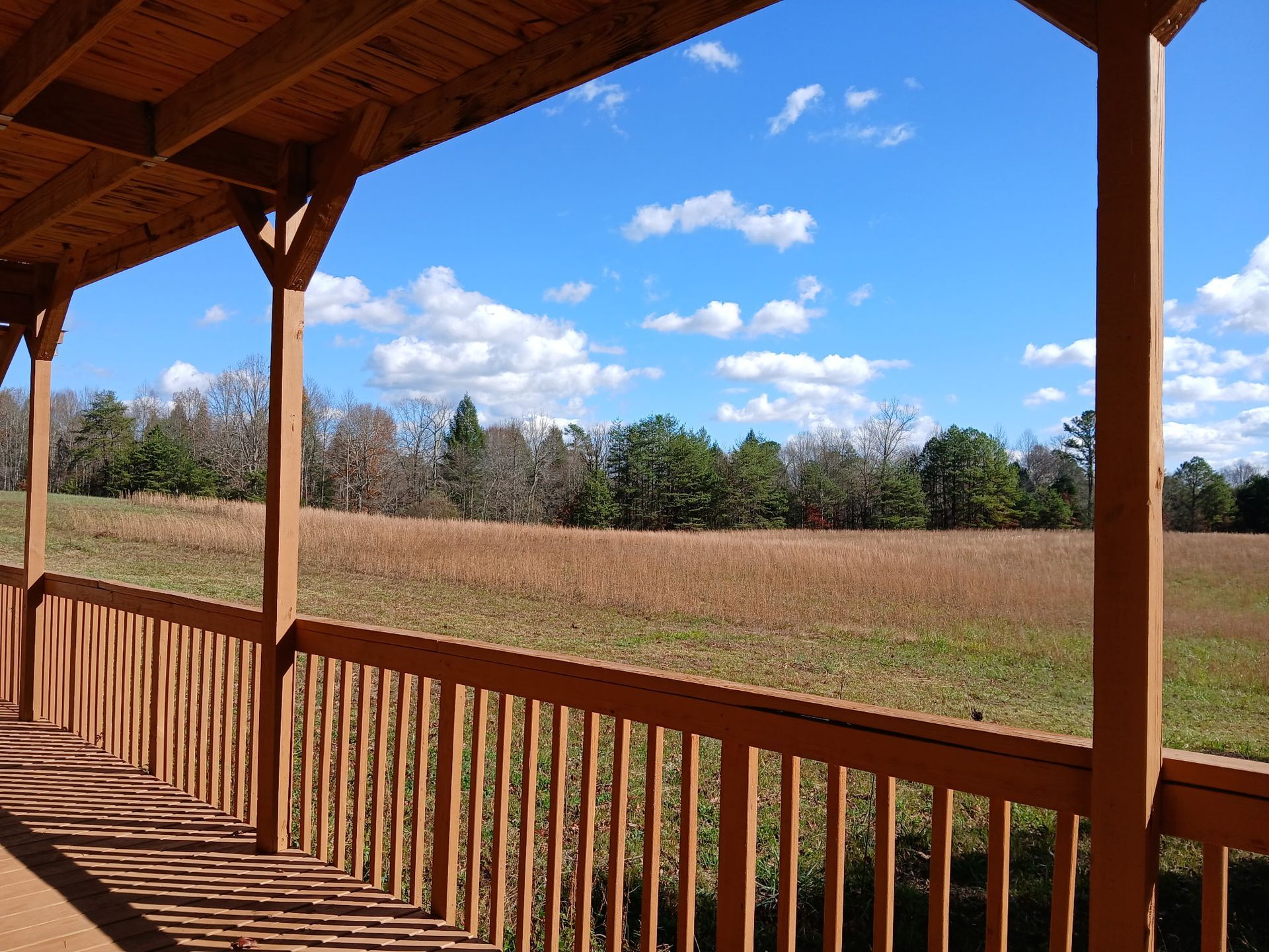 A wooden porch railing looks out over a wide, grassy field toward a line of trees under a bright blue, cloudy sky.