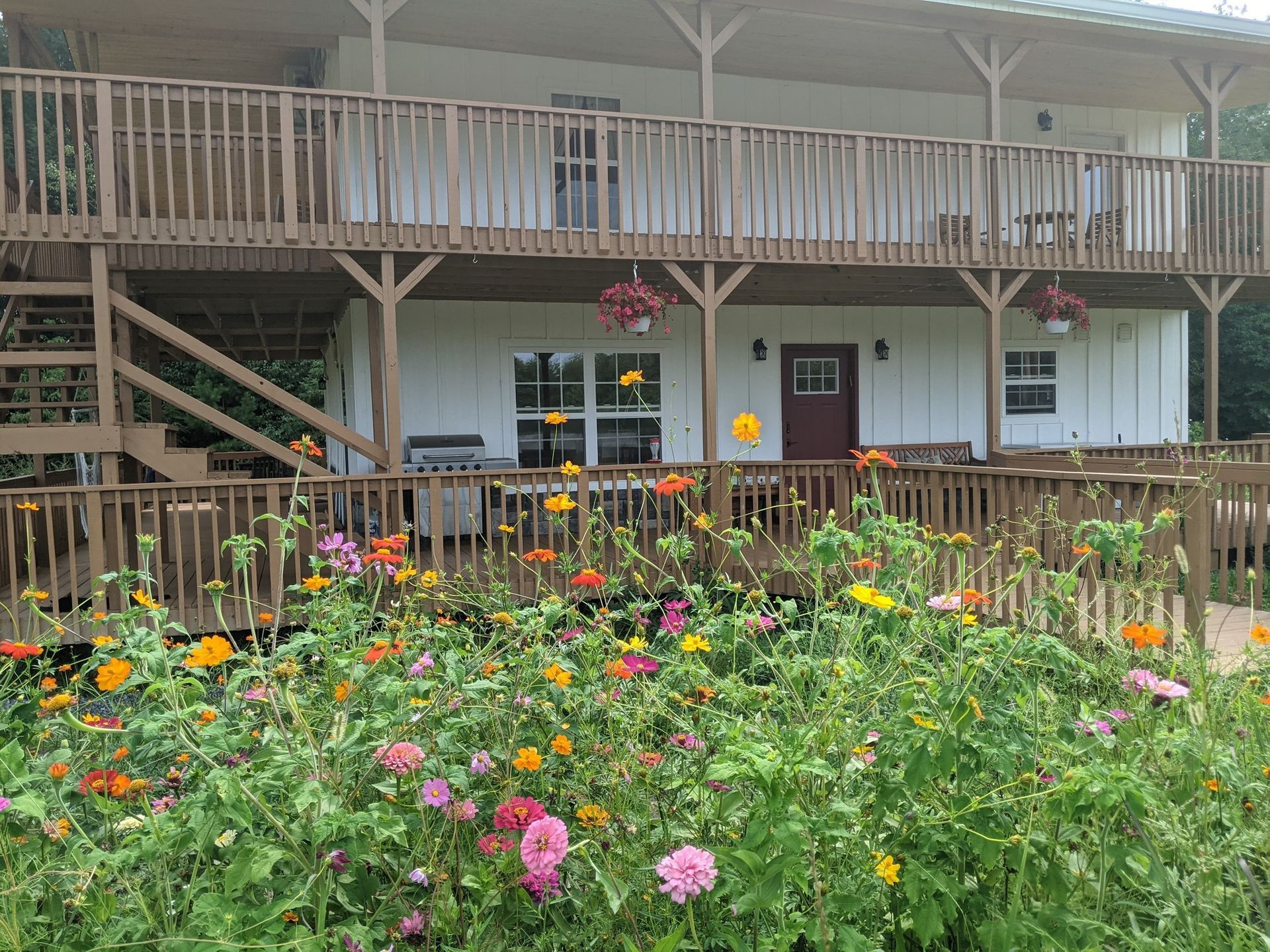 A two-story house with a wooden deck and balcony sits behind a vibrant bed of colorful wildflowers.