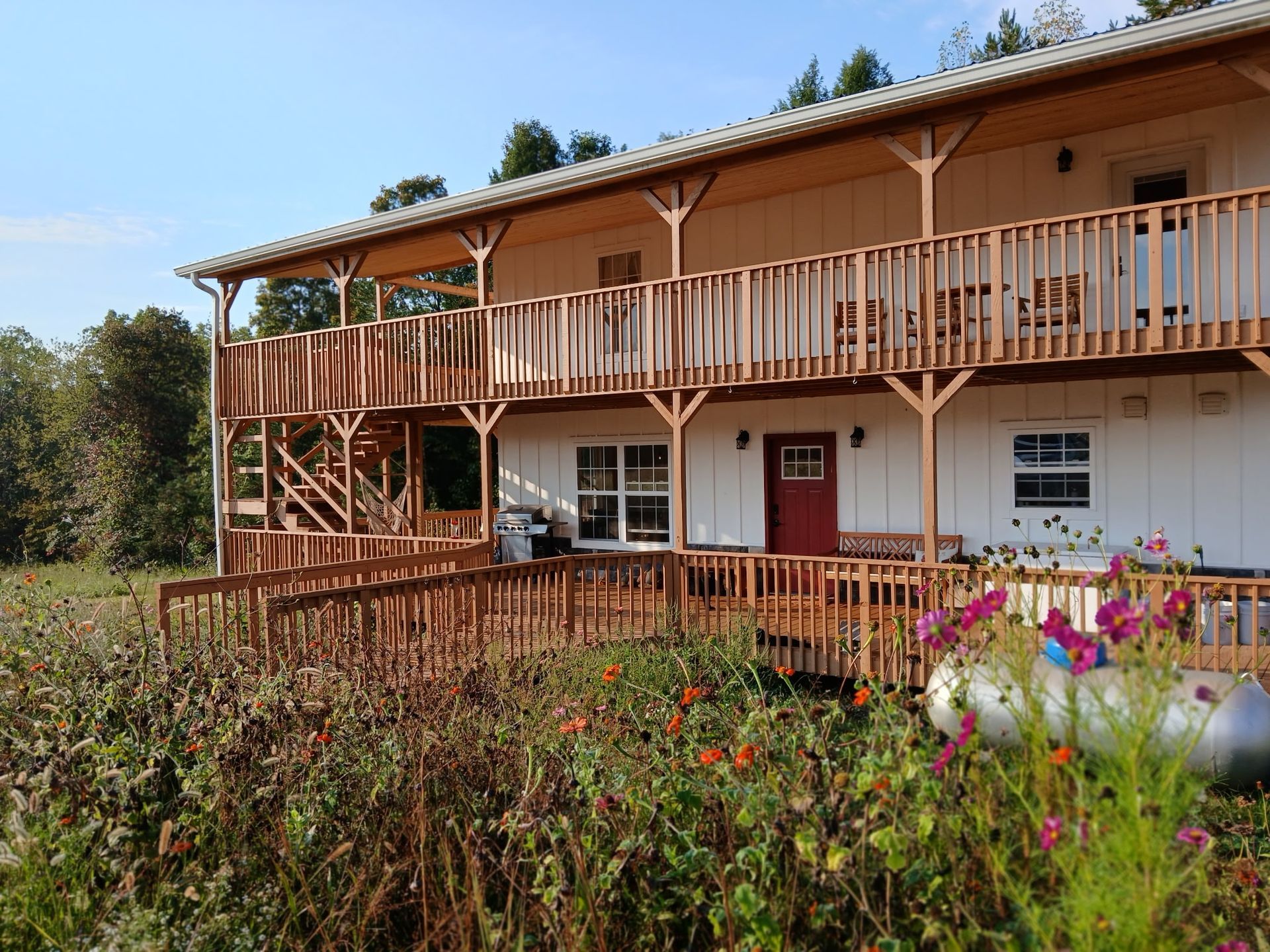 A two-story white building with a wooden deck and balcony, surrounded by greenery and wildflowers on a sunny day.