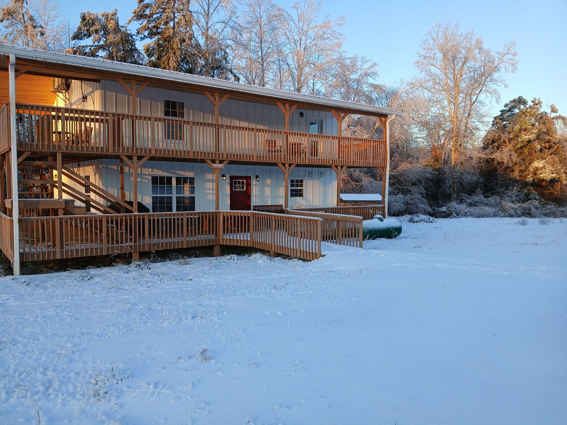 A two-story house with wrap-around wooden decks stands in a snowy, sunlit yard surrounded by frost-covered trees.