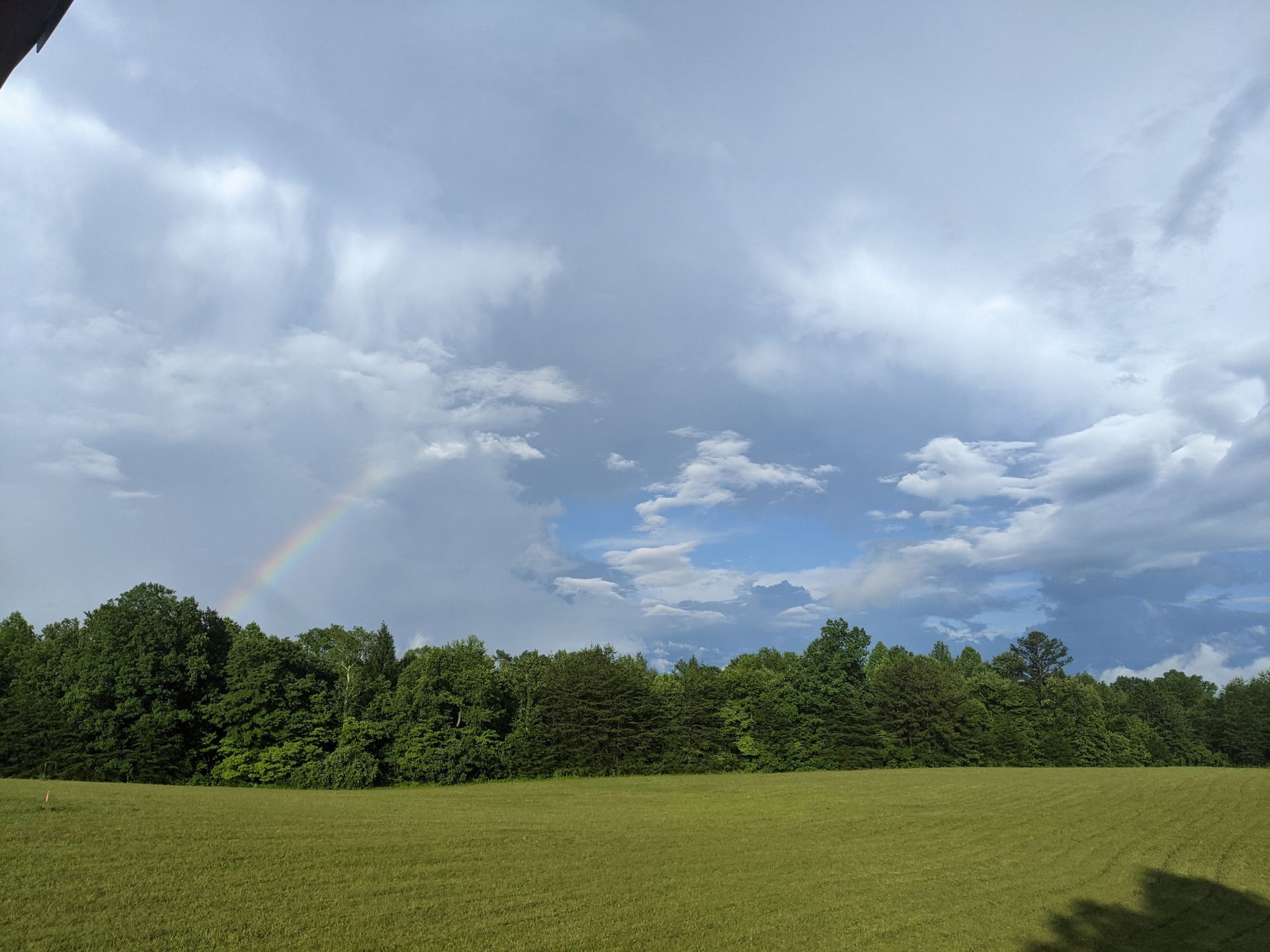 A faint rainbow arches over a dark green tree line above a grassy field under a sky with scattered clouds.