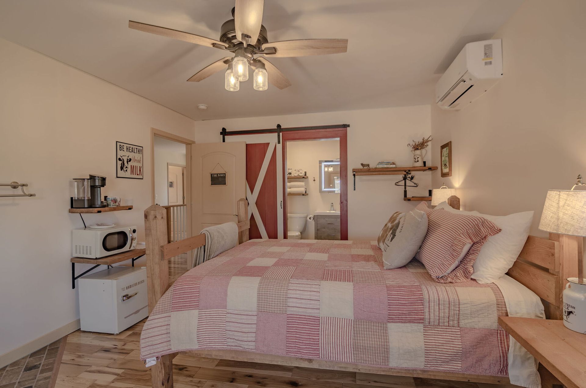 A bedroom with a queen bed, a quilt, a ceiling fan, a microwave, a small fridge, and a bathroom with a sliding barn door.
