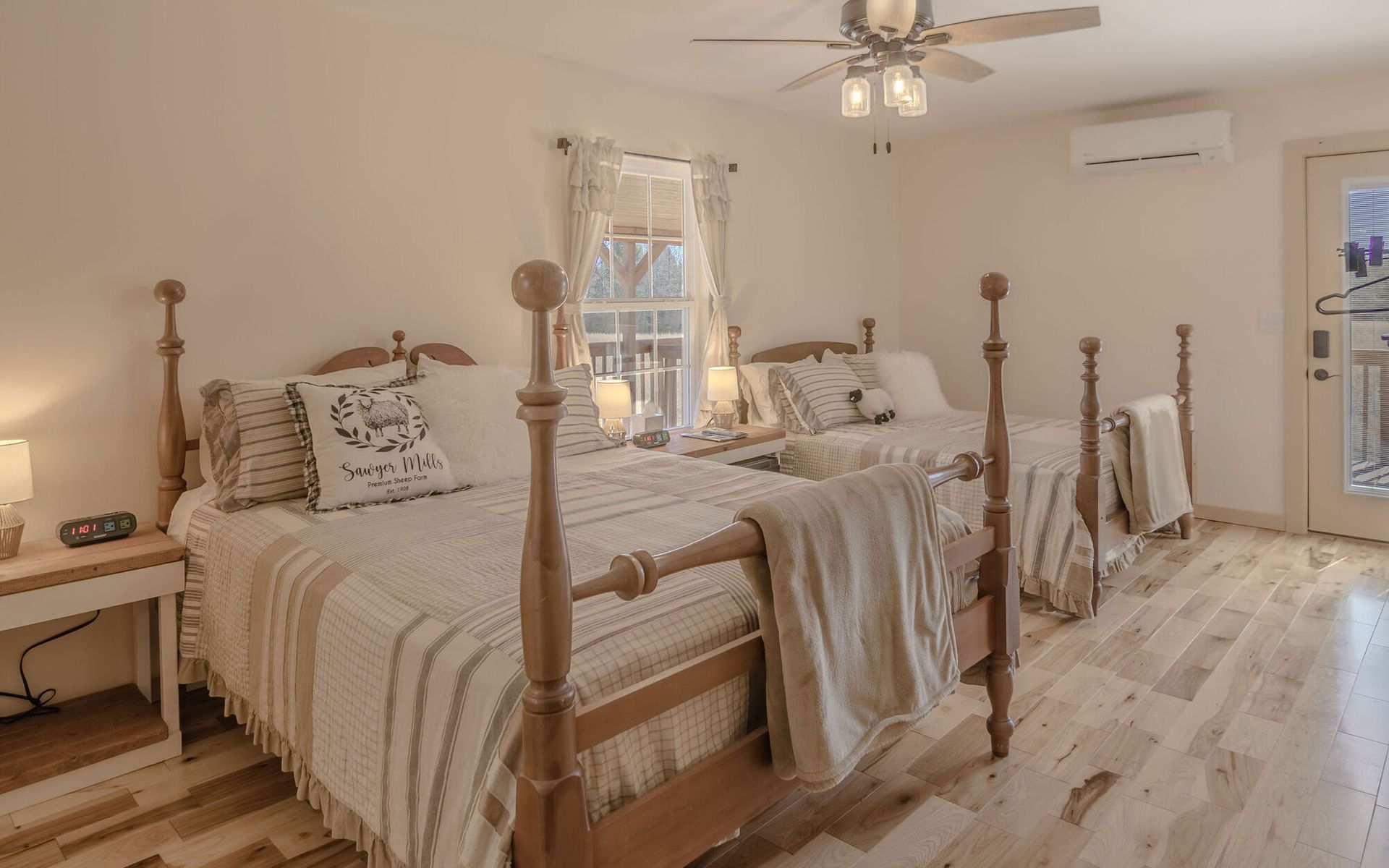 A bedroom with two wooden post beds, neutral bedding, light wood floors, a ceiling fan, and a door to a balcony.