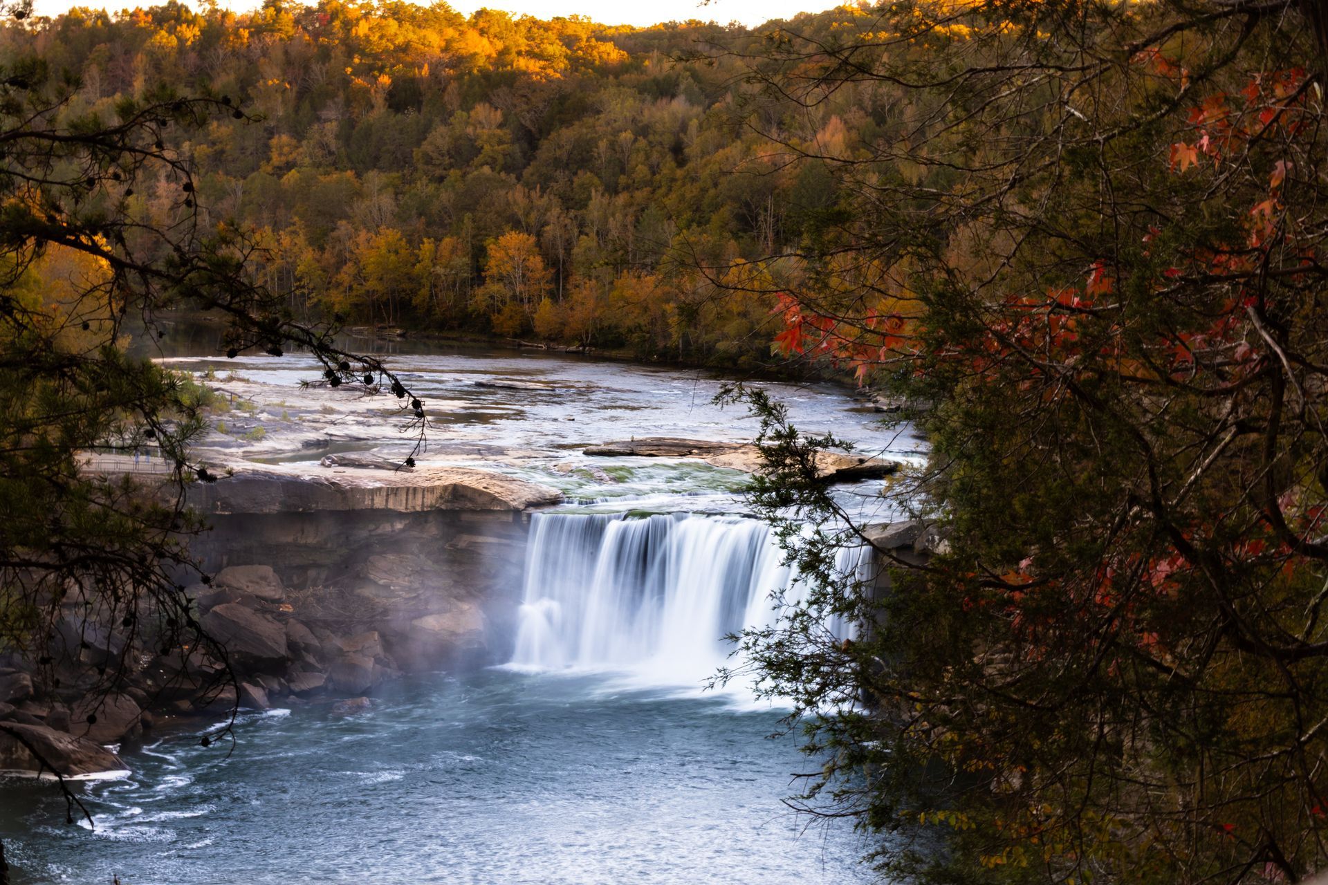 A waterfall flows over a rocky ledge into a river, surrounded by trees with autumn foliage under warm, golden light.
