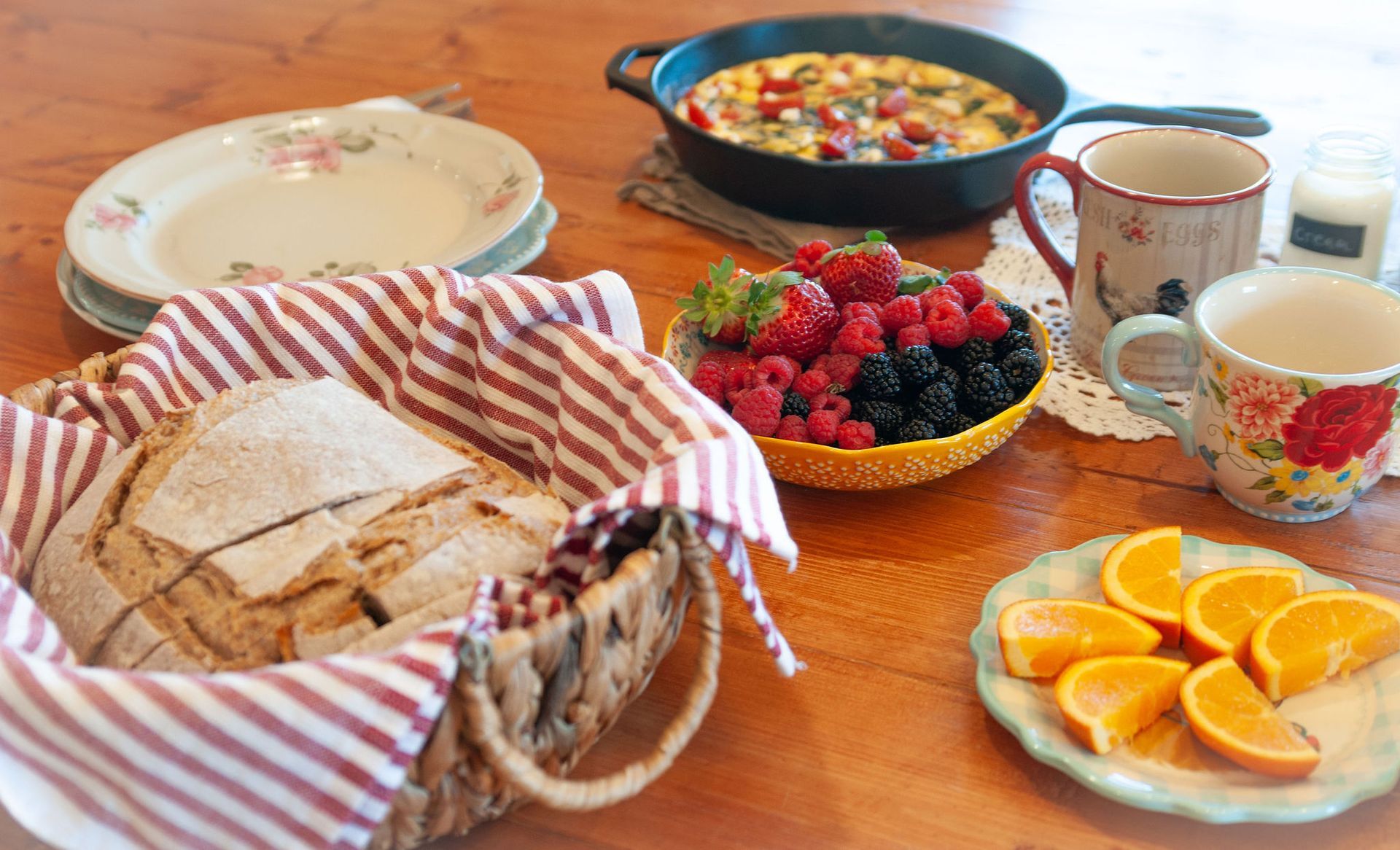 A table setting featuring a loaf of bread in a basket, a skillet of food, a bowl of berries, and slices of orange.