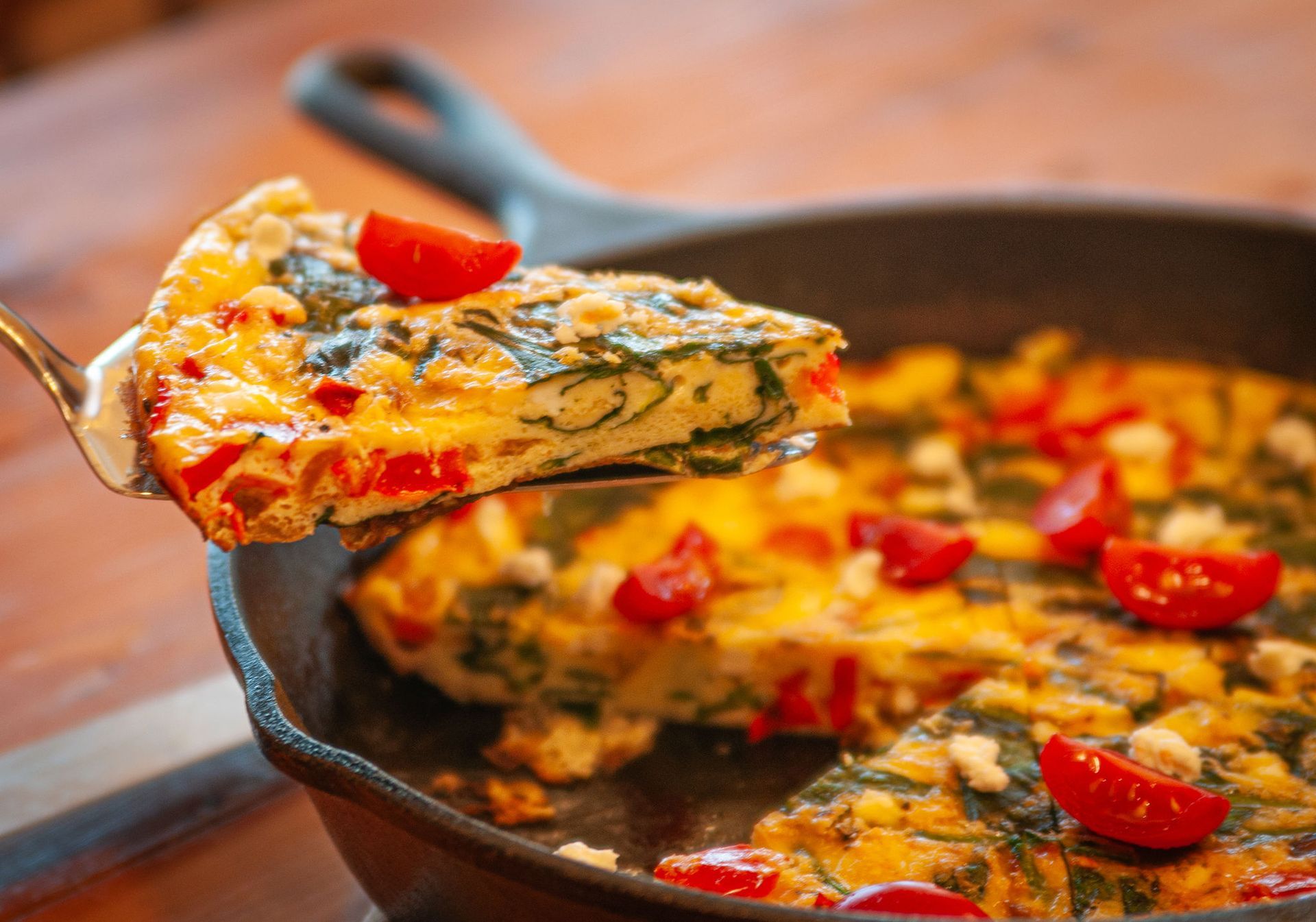 A slice of vegetable frittata with tomatoes and spinach, lifted by a spatula from a black cast-iron skillet.