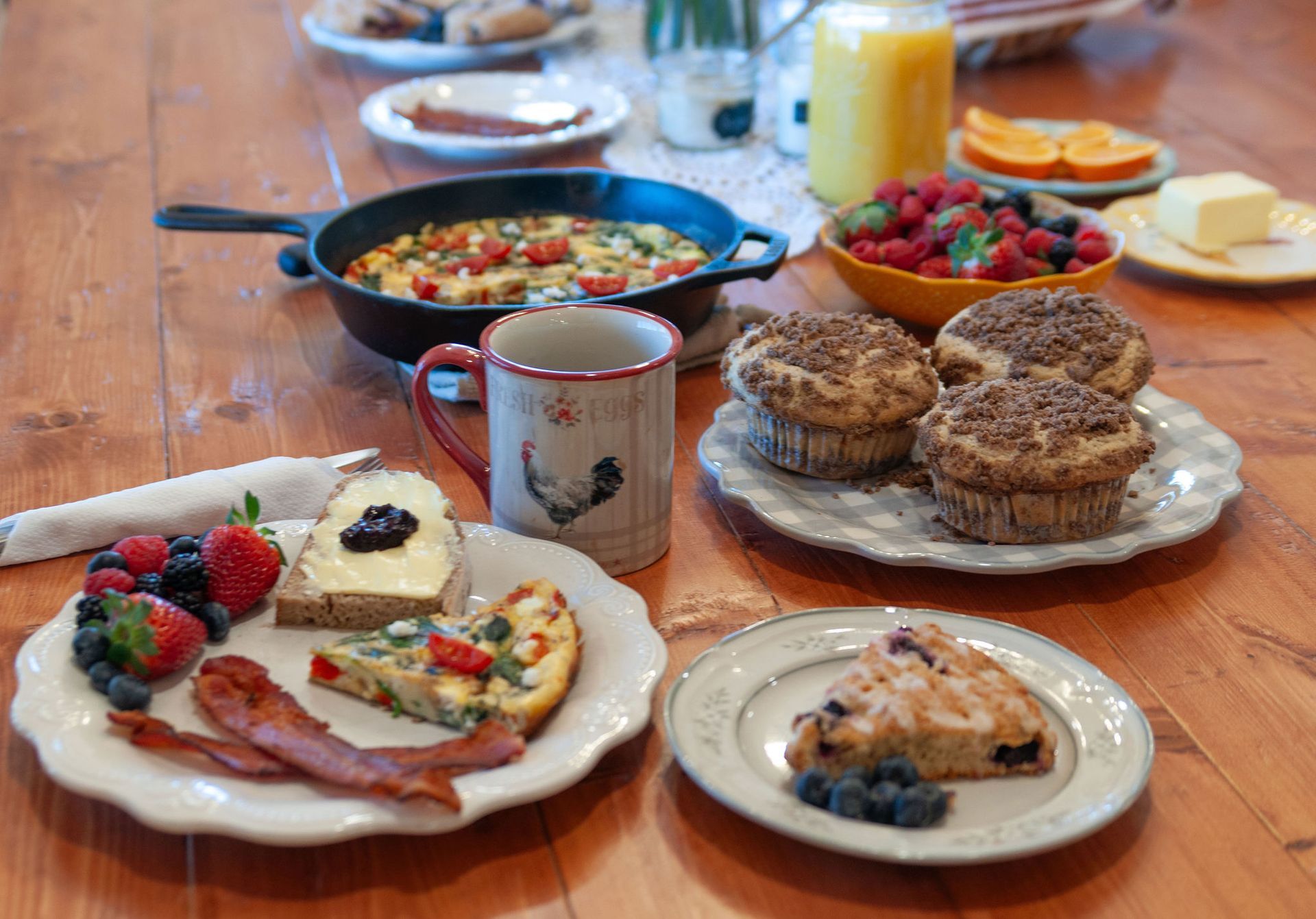 A rustic wooden table set with a breakfast feast, including a frittata, muffins, bacon, fruit, butter, and coffee.