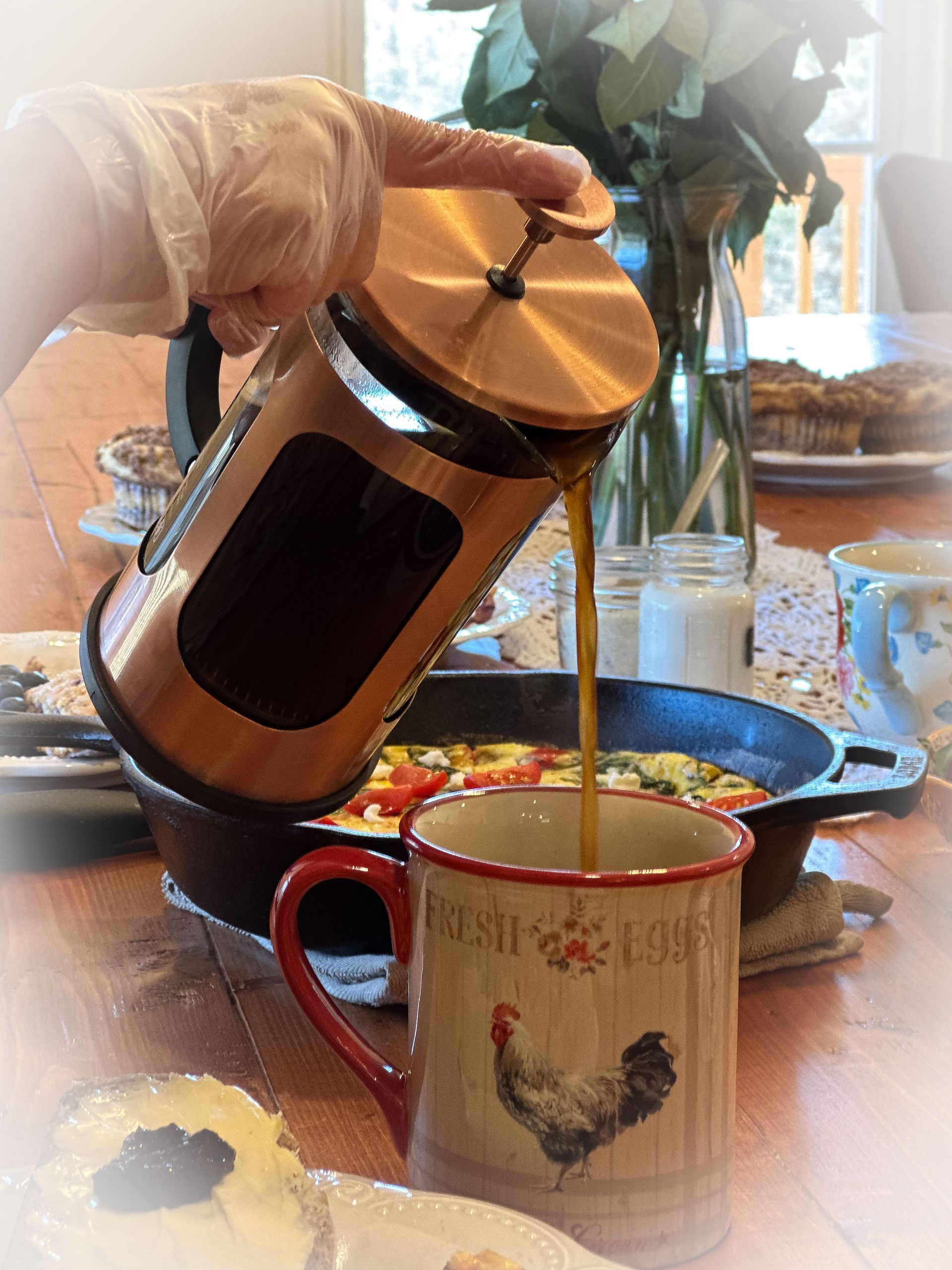 A person pours coffee from a copper-trimmed French press into a rooster-patterned mug on a rustic table with a skillet.