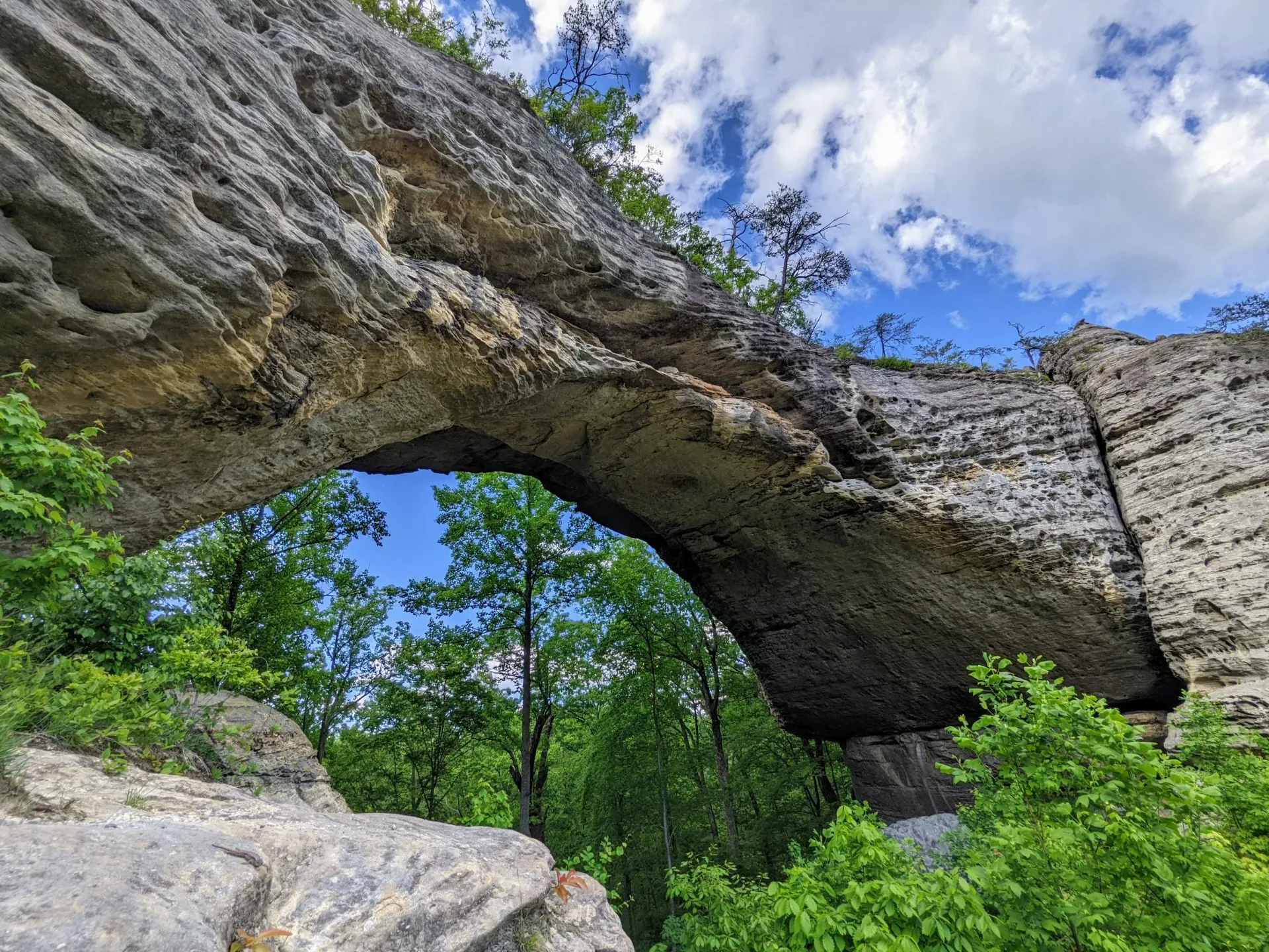A natural rock arch spans across a bright blue sky, surrounded by lush green trees and rocky cliffs.